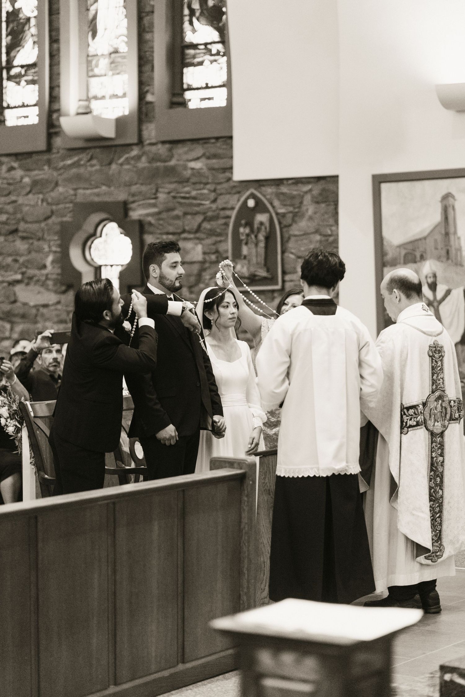 Priest places veil over bride and groom during traditional Catholic wedding ceremony at church altar.