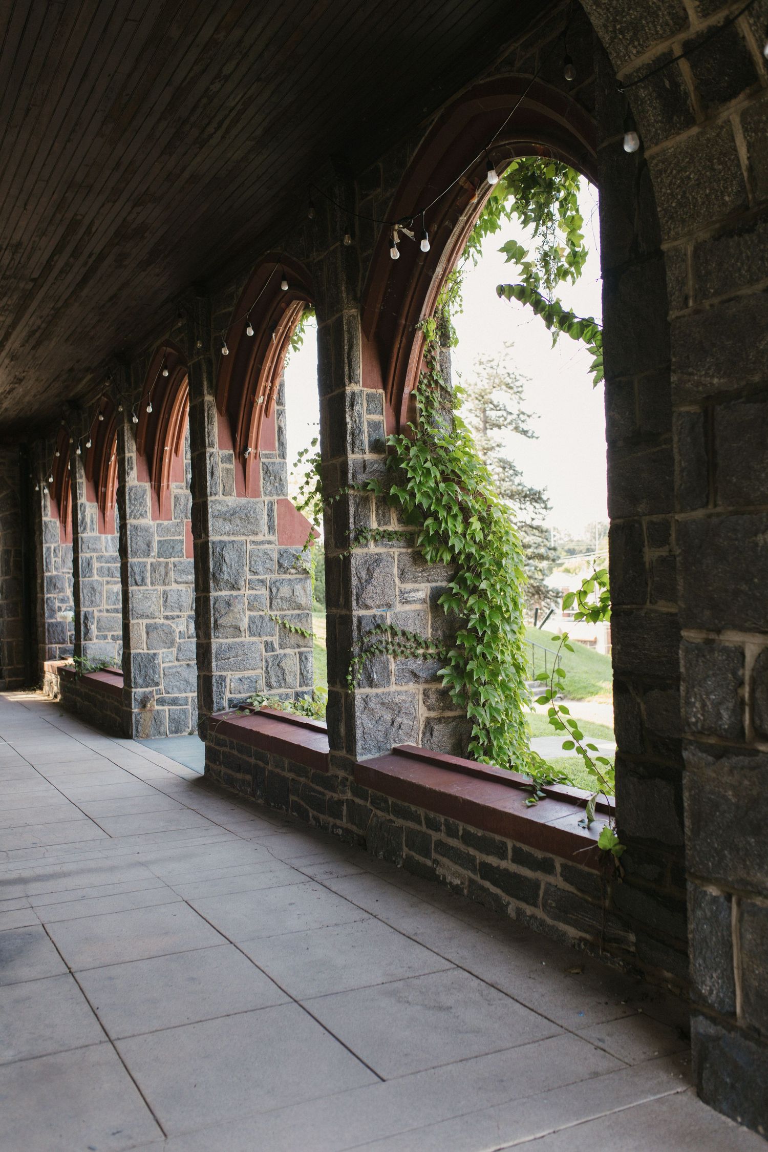 Gothic stone church corridor with arched openings, ivy-covered walls, and a tiled walkway leading through elegant architectural arches.