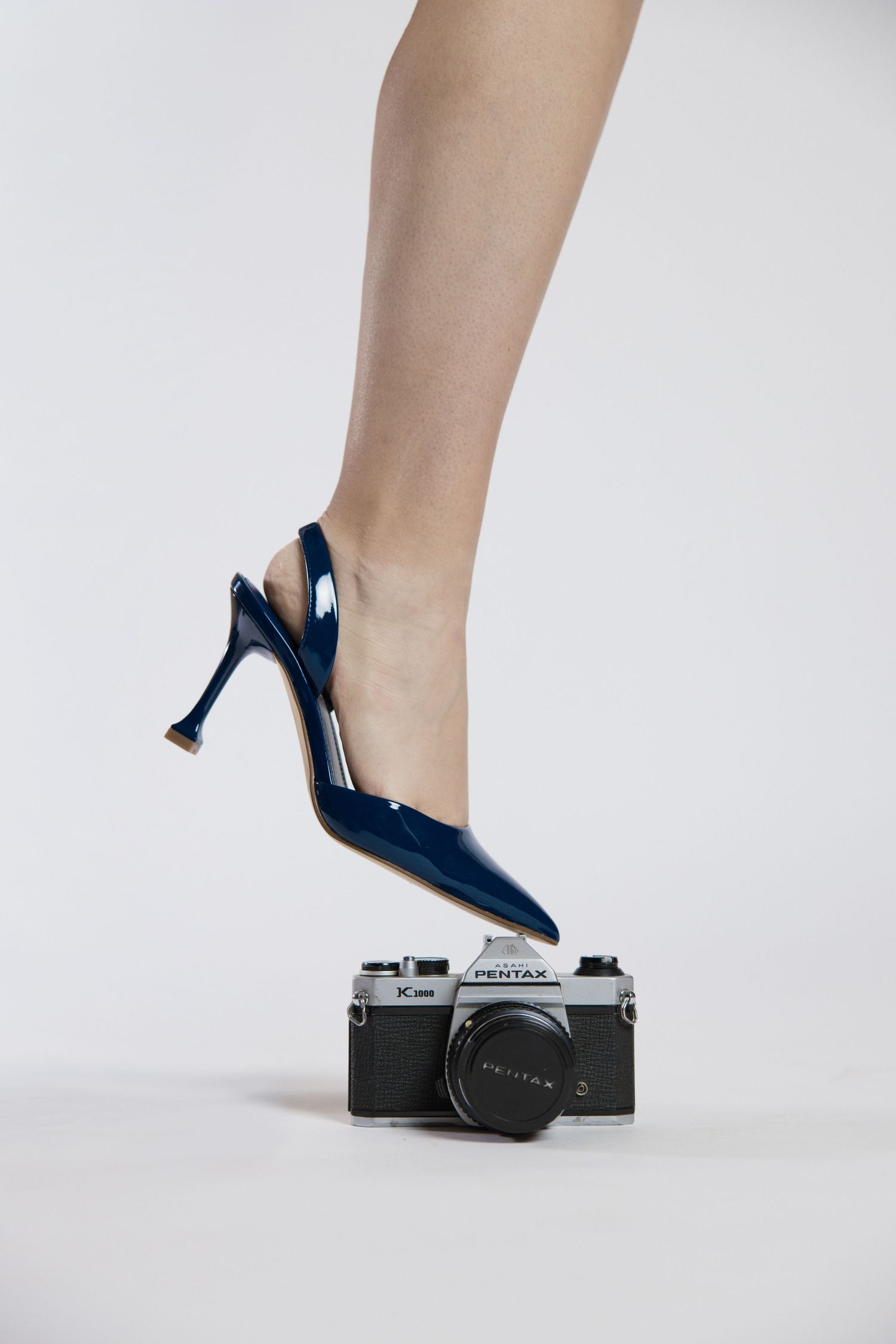 A woman's leg in a navy blue patent slingback stiletto heel stepping on a vintage Pentax film camera.