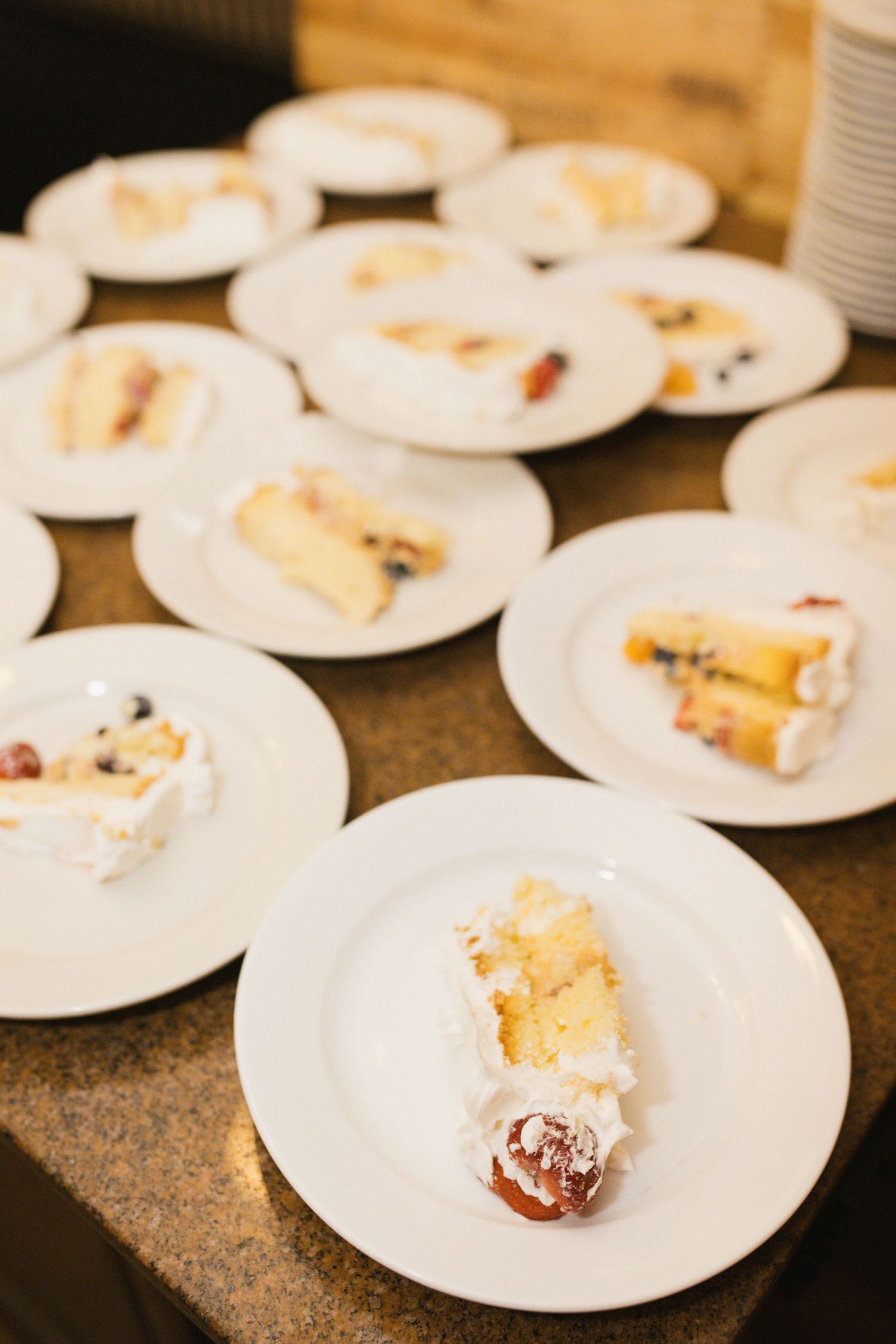 Multiple slices of white layered wedding cake with fruit served on white plates at reception.