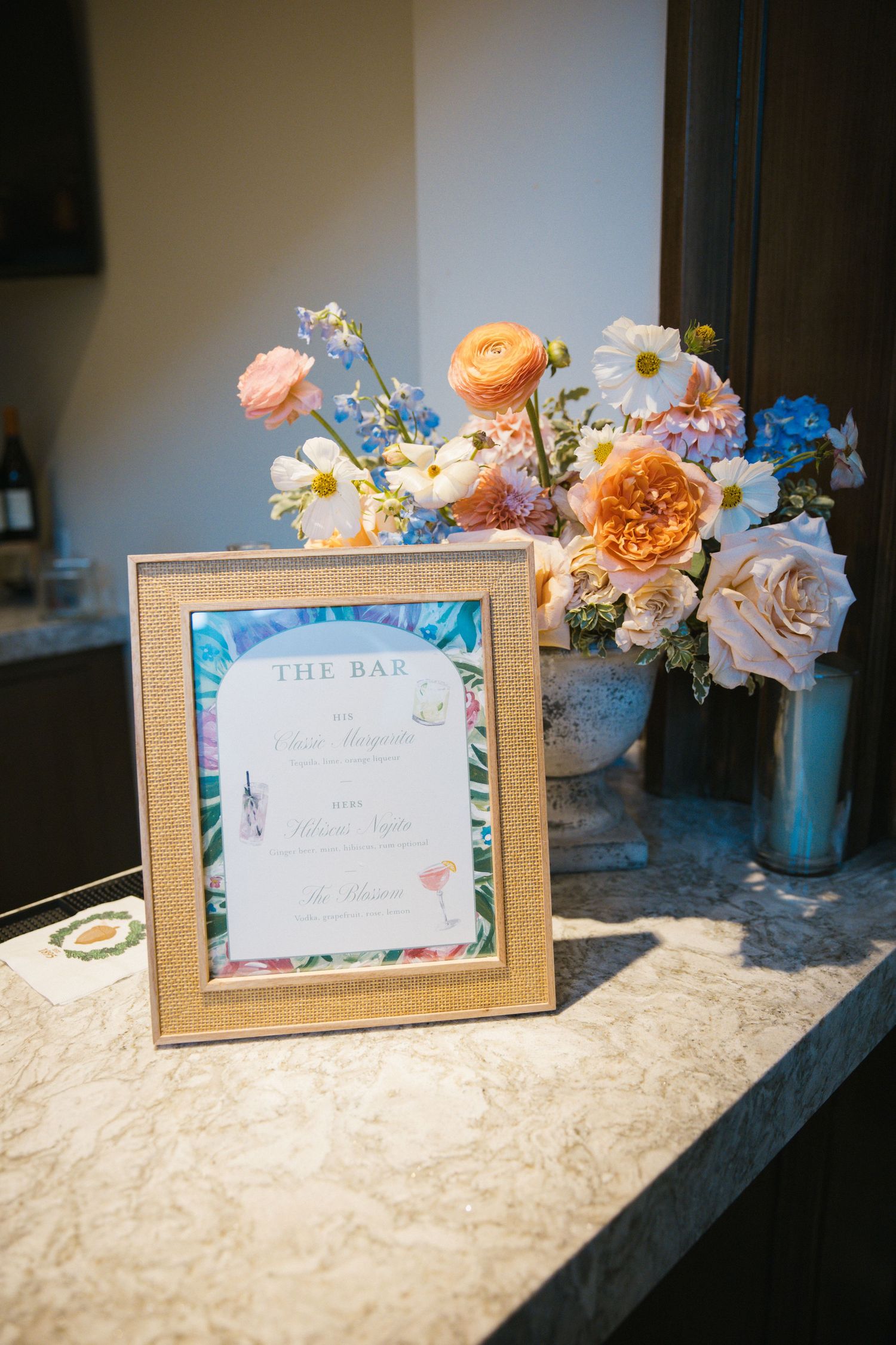 Gold-framed bar menu sign beside a vase of peach, white, and blue flowers on marble counter.