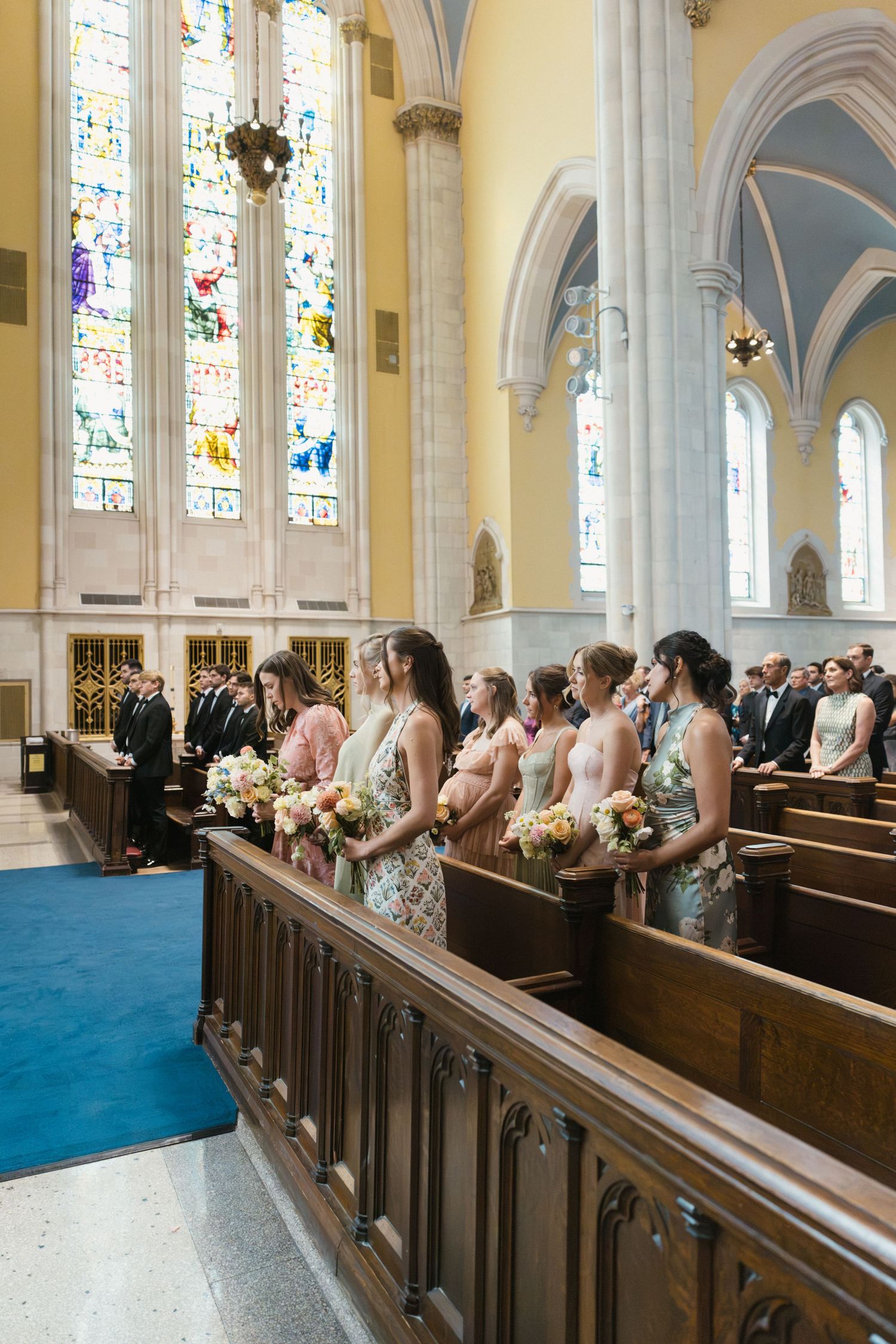 Bridesmaids in pastel dresses holding floral bouquets stand in church pews during a wedding ceremony.