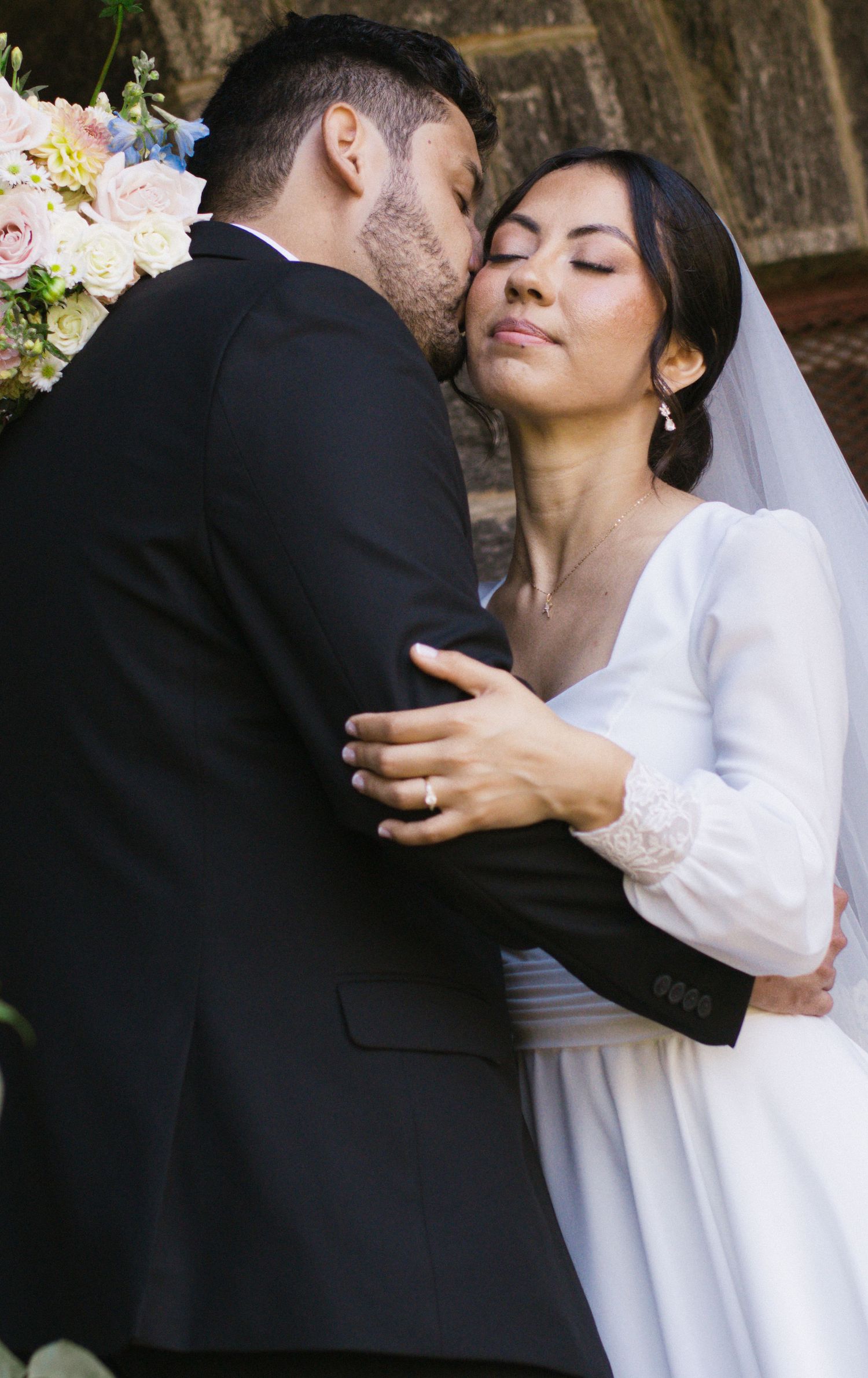 A groom in a black suit kisses his bride in a white gown and veil during their wedding ceremony.