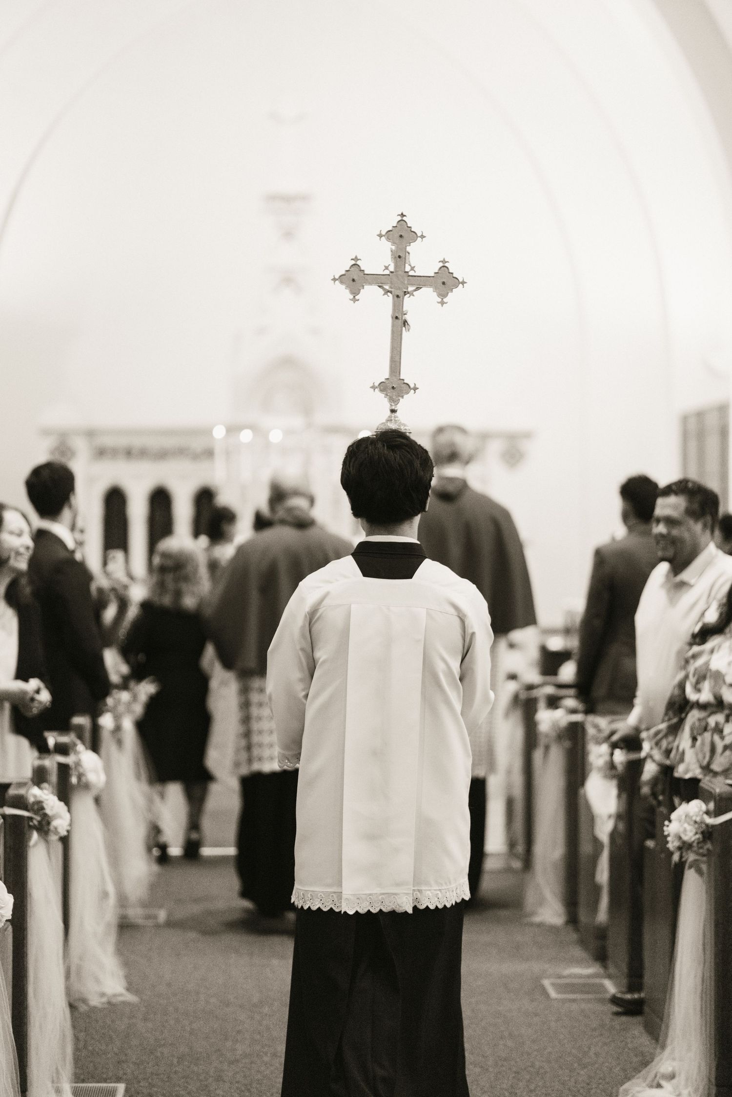 Altar server carrying ornate cross down church aisle during wedding processional, black and white.