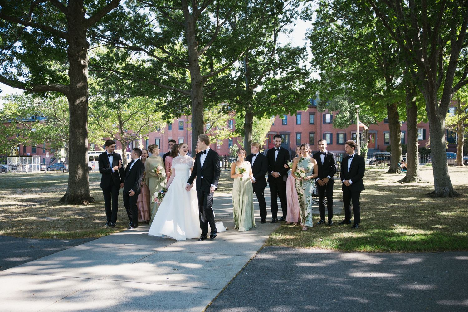 Bride and groom with wedding party gathered outdoors under lush green trees on a sunny day.