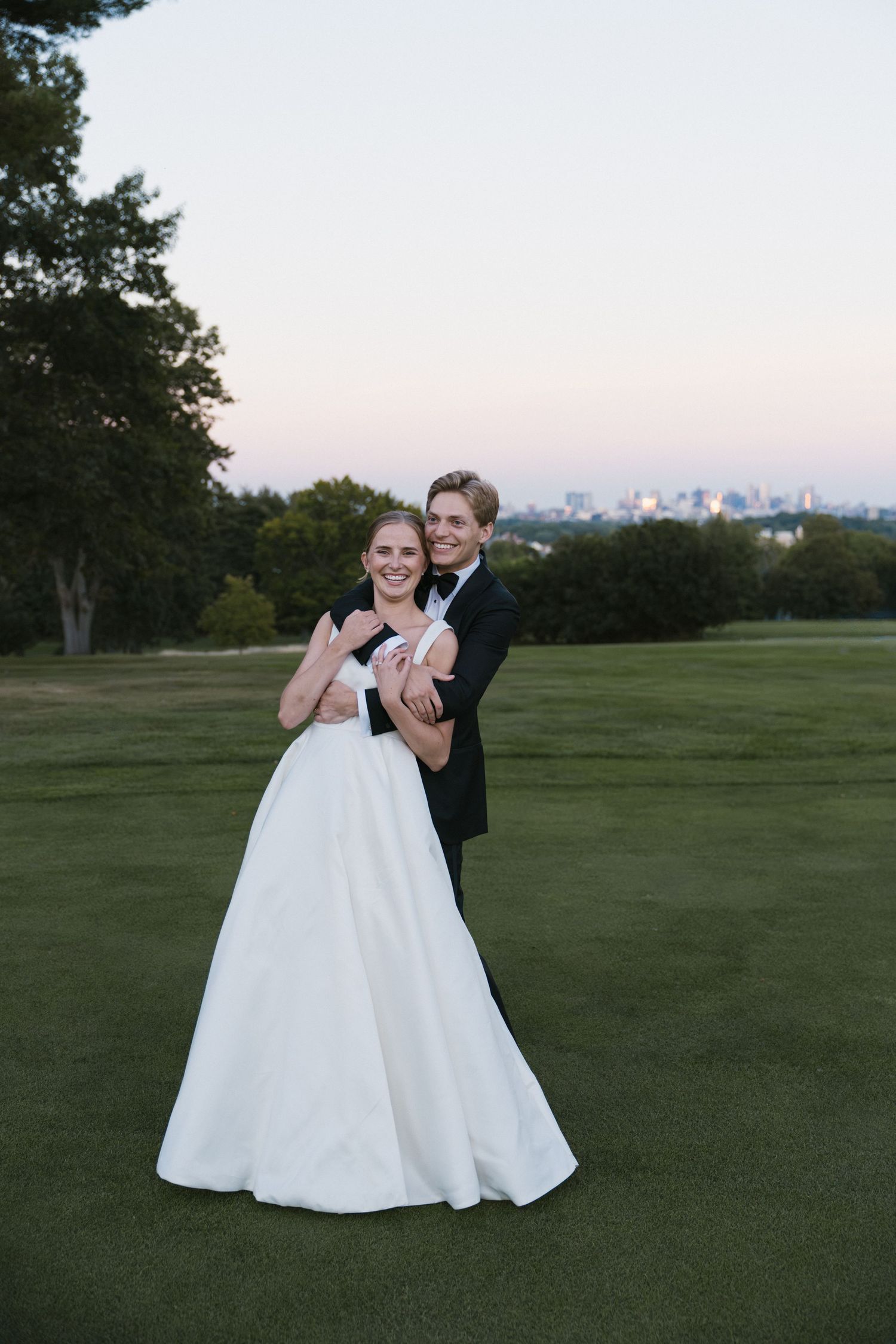 Bride and groom embracing on golf course lawn at dusk with city skyline visible in background.