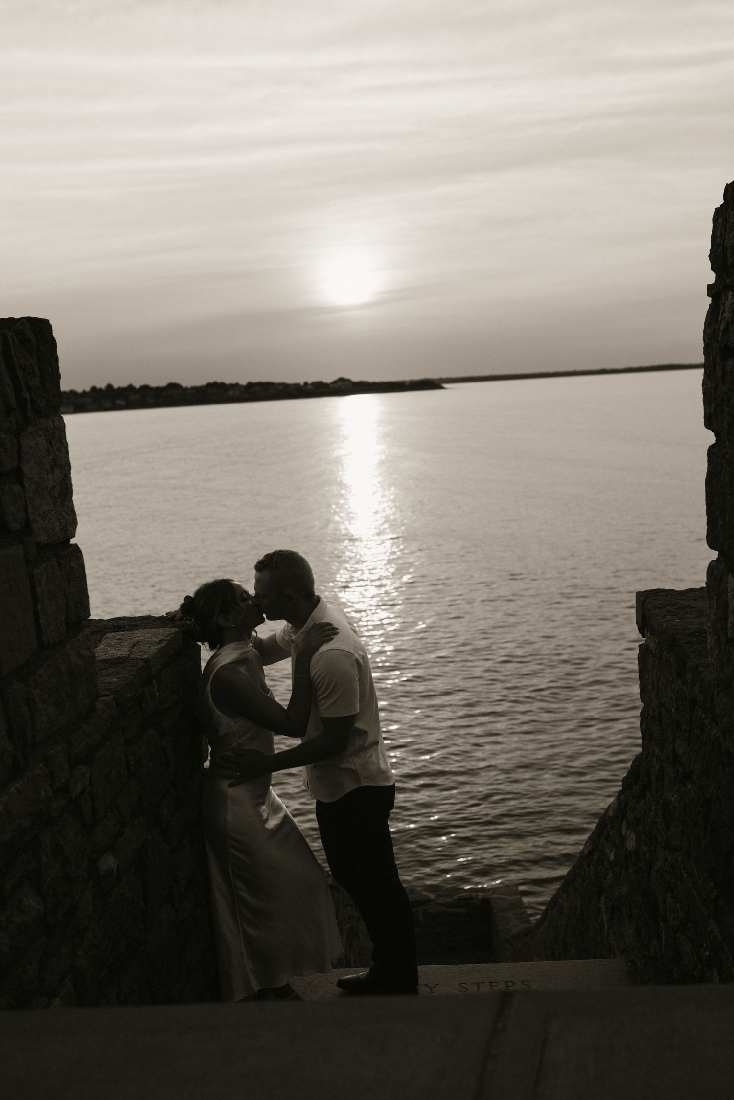 Black and white photo of couple kissing at sunset between stone pillars by the sea.