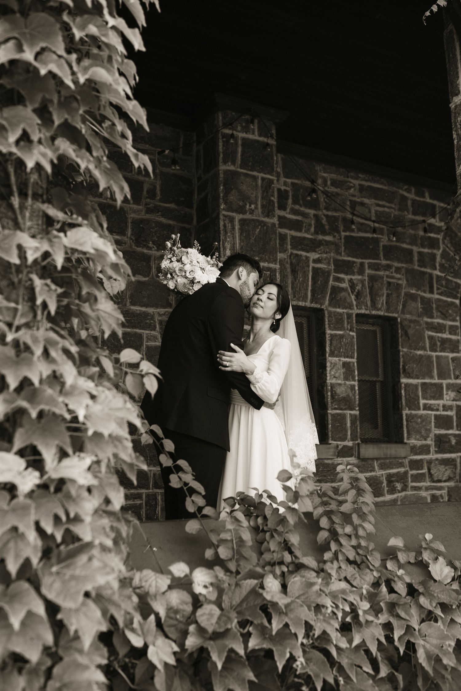 Black and white photo of bride and groom embracing on ivy-covered stone church steps.