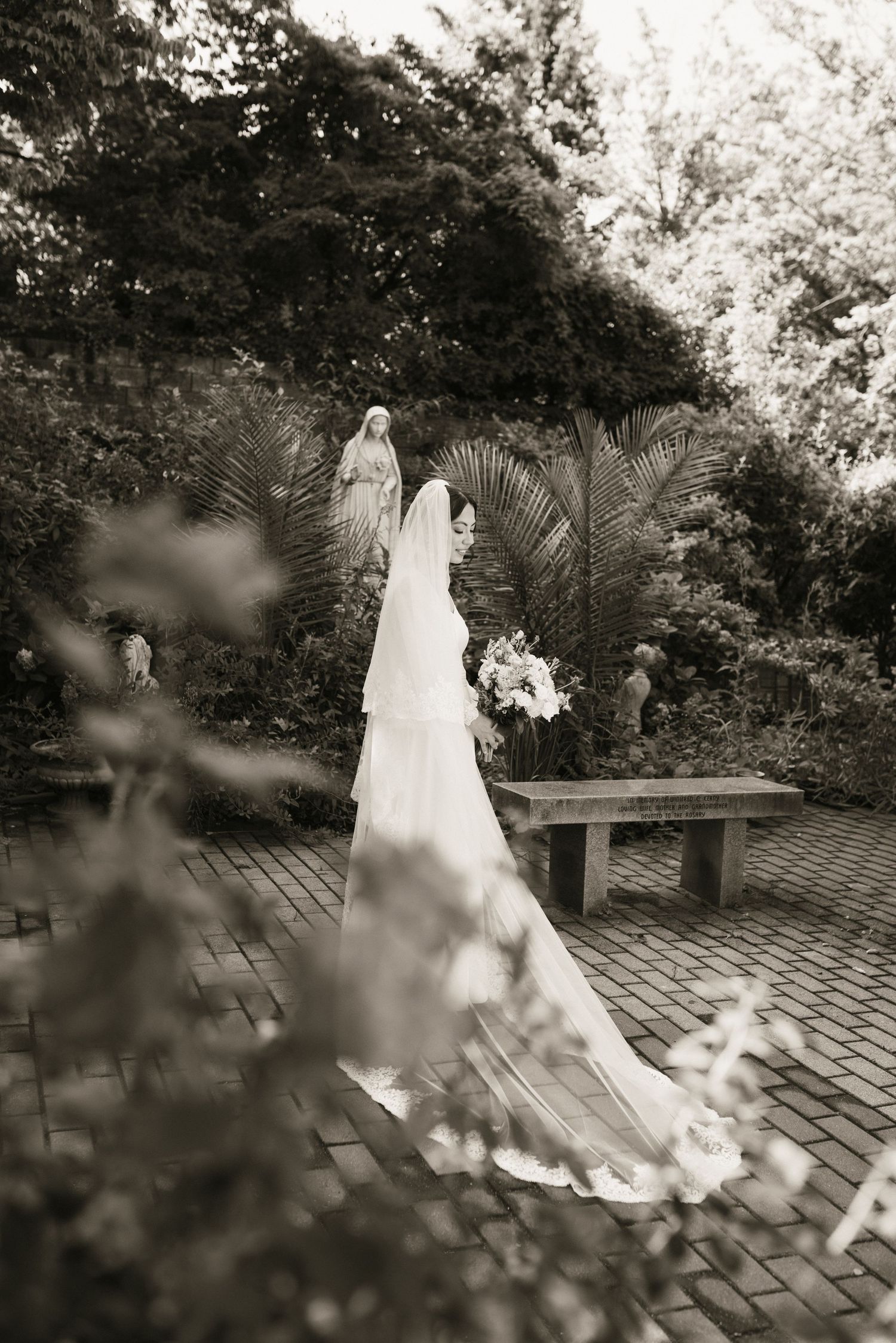 Bride in flowing white gown and cathedral veil stands in lush garden near statue, holding bouquet in black and white photo.