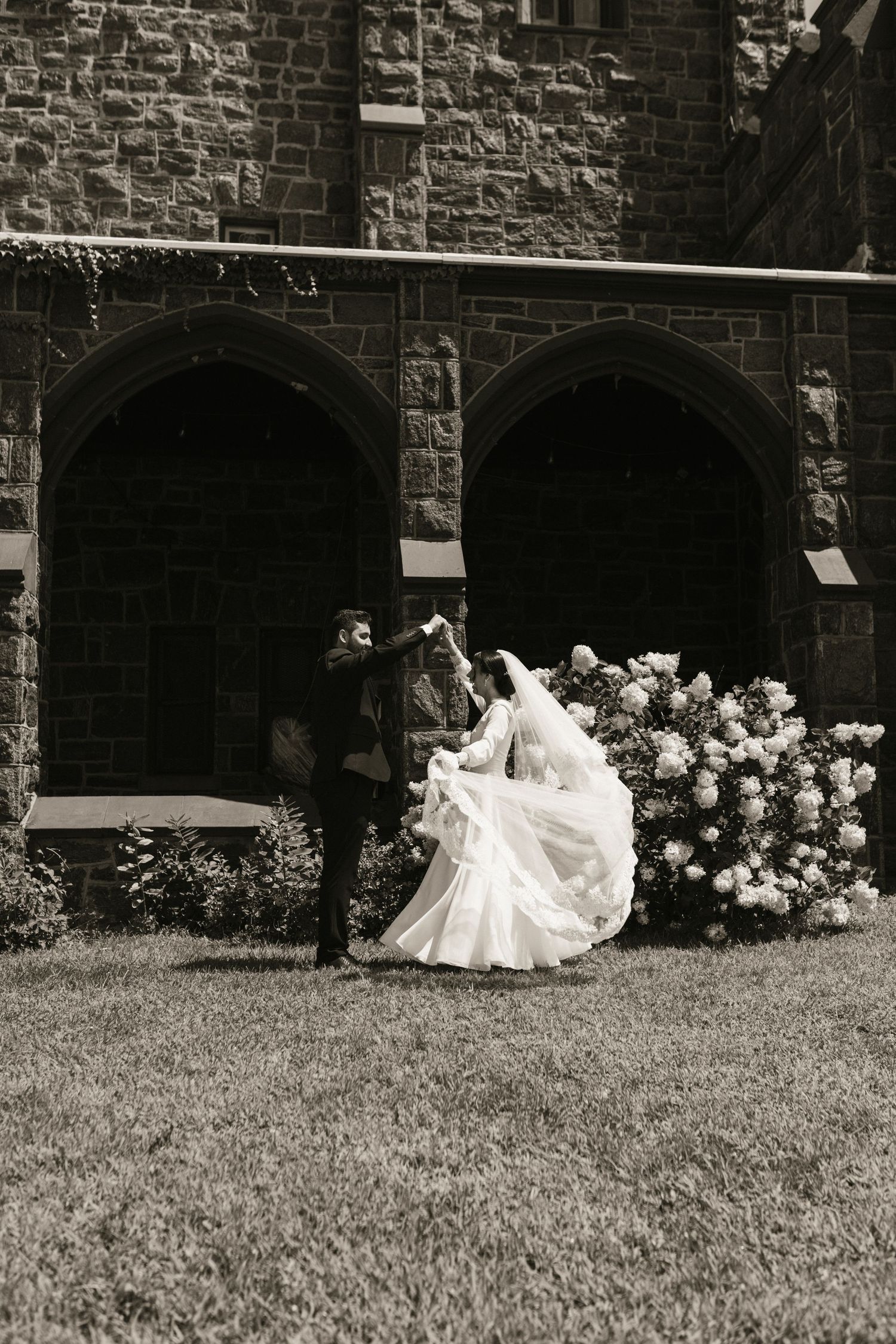 Bride and groom dance outside historic stone church with Gothic arches and blooming white floral bushes in black and white.