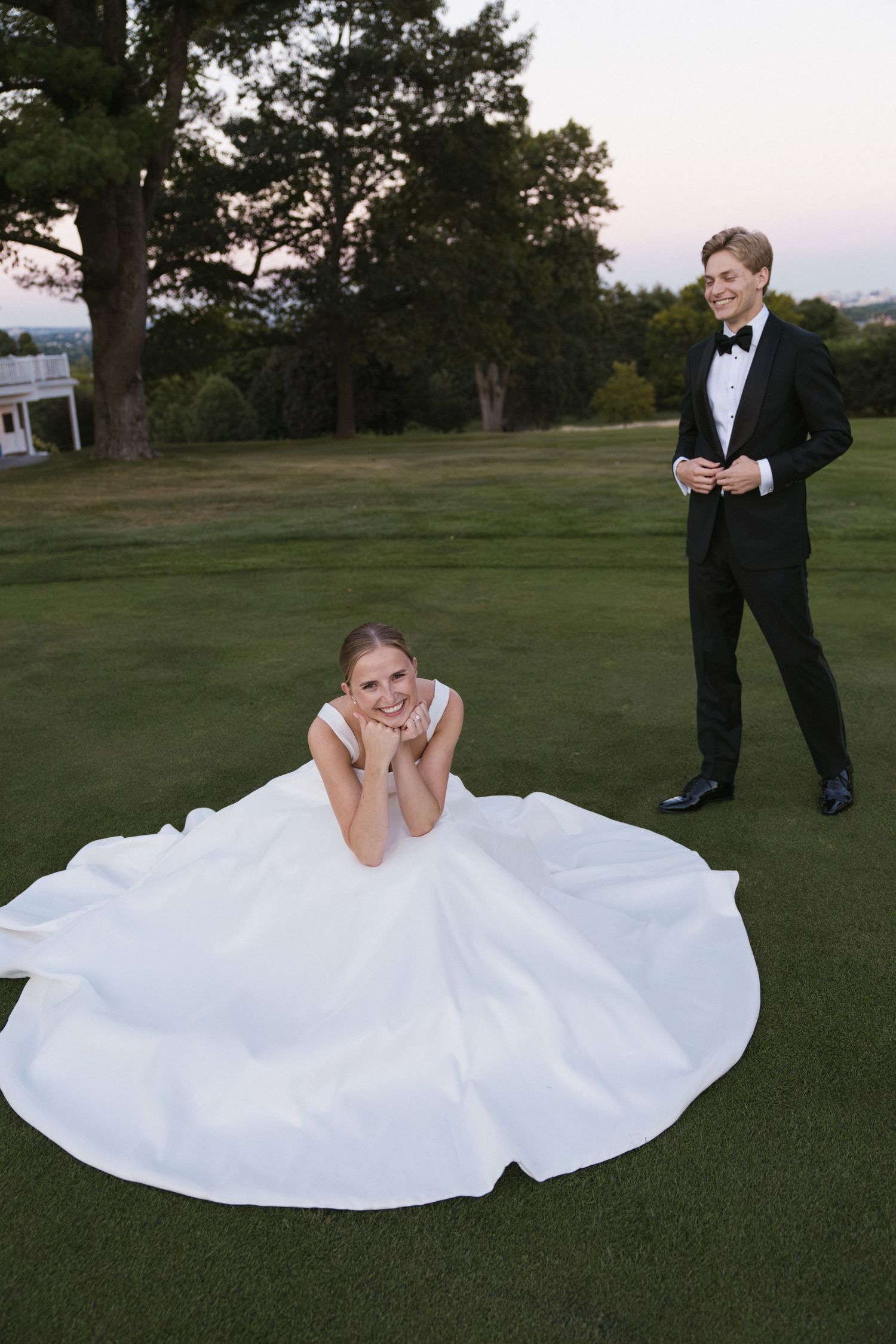 Bride in a white ball gown poses on grass while groom in black tuxedo stands smiling behind her.
