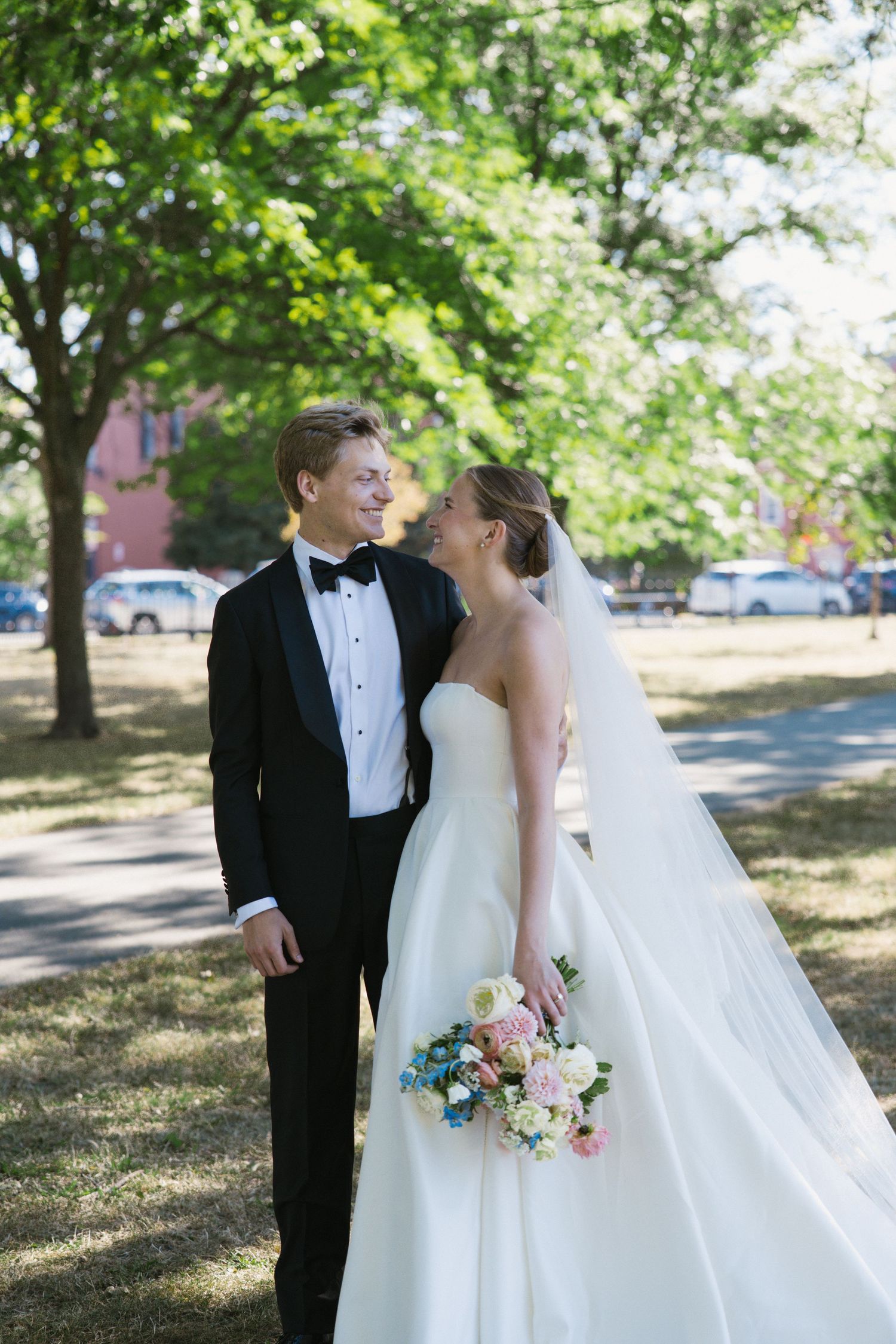Bride and groom share a romantic moment outdoors under green trees, bride holding a pastel bouquet in a strapless gown.