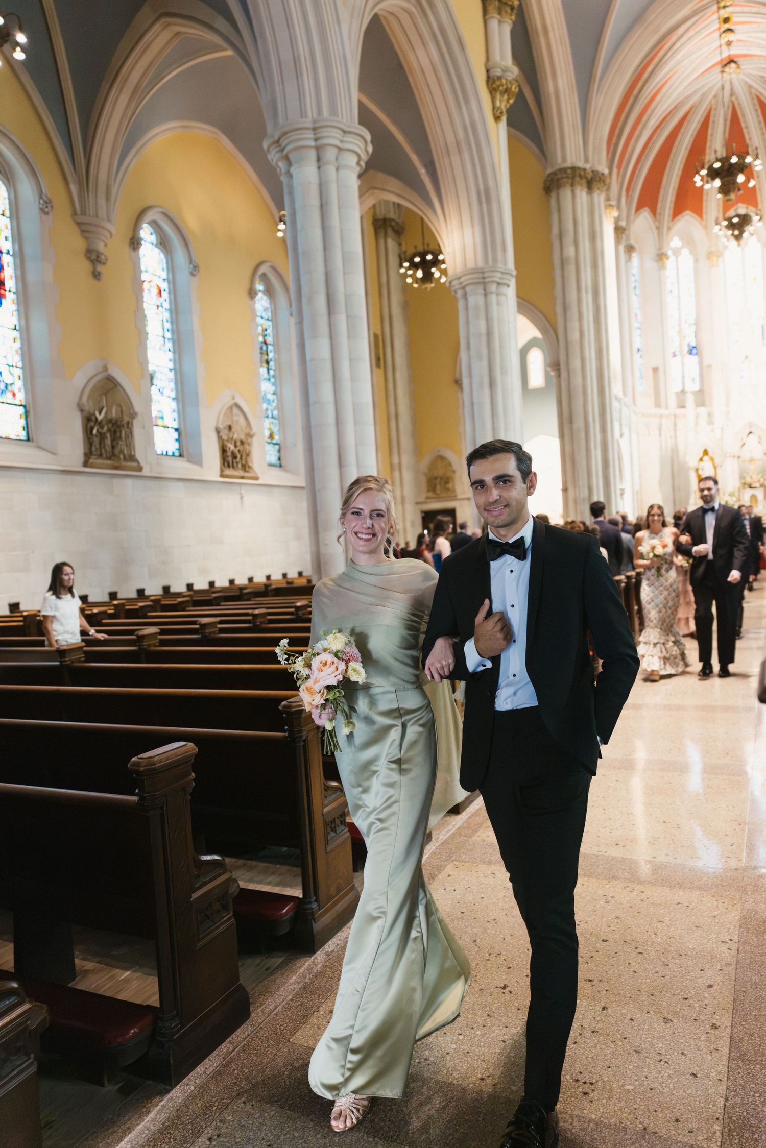 Bride in sage green gown walks arm-in-arm with groomsman in black tuxedo down church aisle.