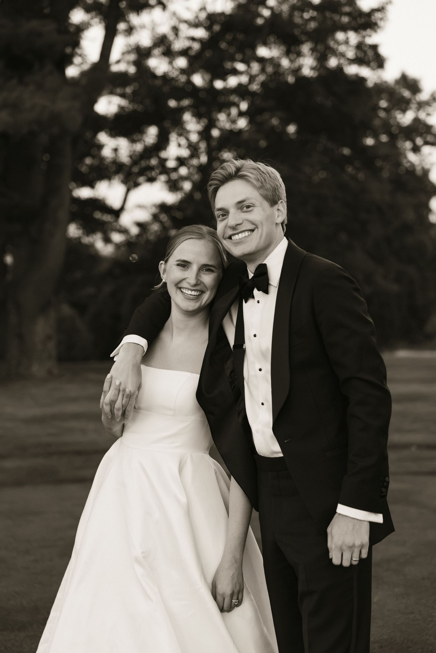 Black and white portrait of smiling bride and groom in tuxedo posing outdoors at wedding.