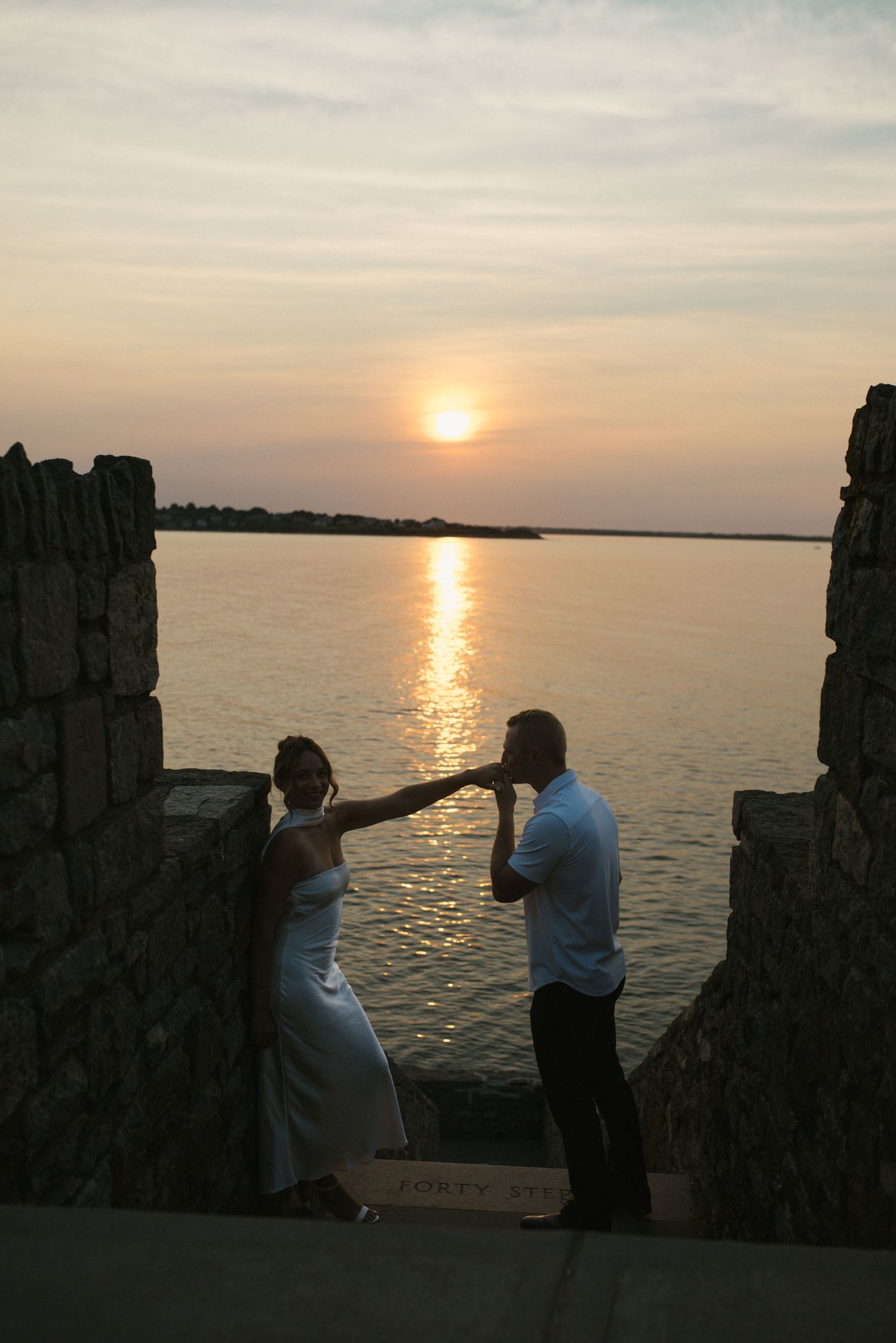 Couple dancing at sunset by stone ruins overlooking calm ocean waters with golden sun reflection.