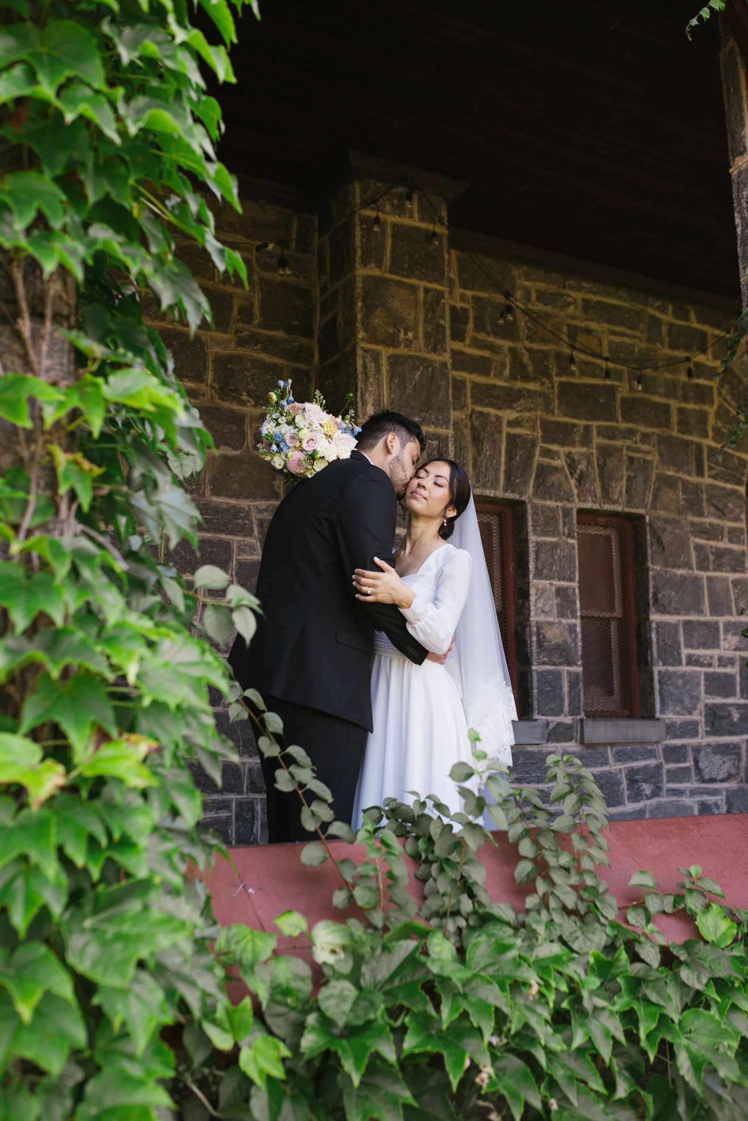 Bride and groom embrace on stone church steps surrounded by lush green ivy.