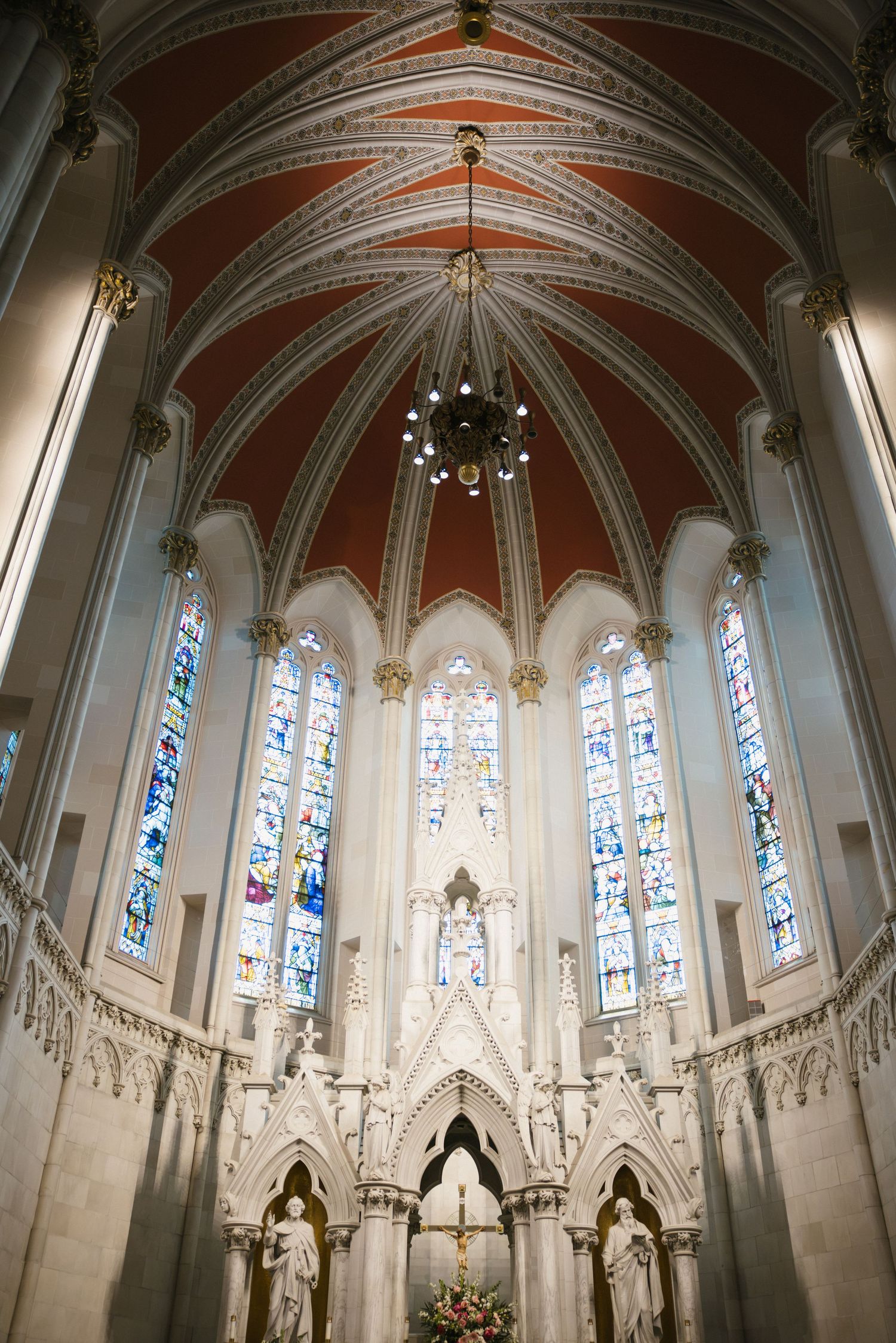 Gothic cathedral interior with ornate white altar, colorful stained glass windows, and ribbed vaulted ceiling painted in red.