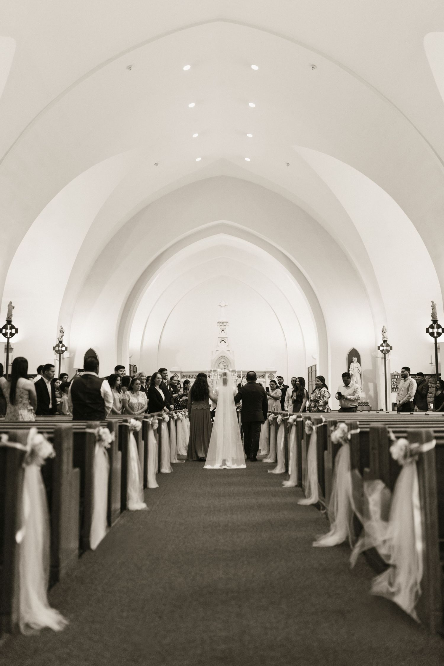 Black and white photo of a wedding ceremony in a church with arched ceilings, pews adorned with white ribbons.