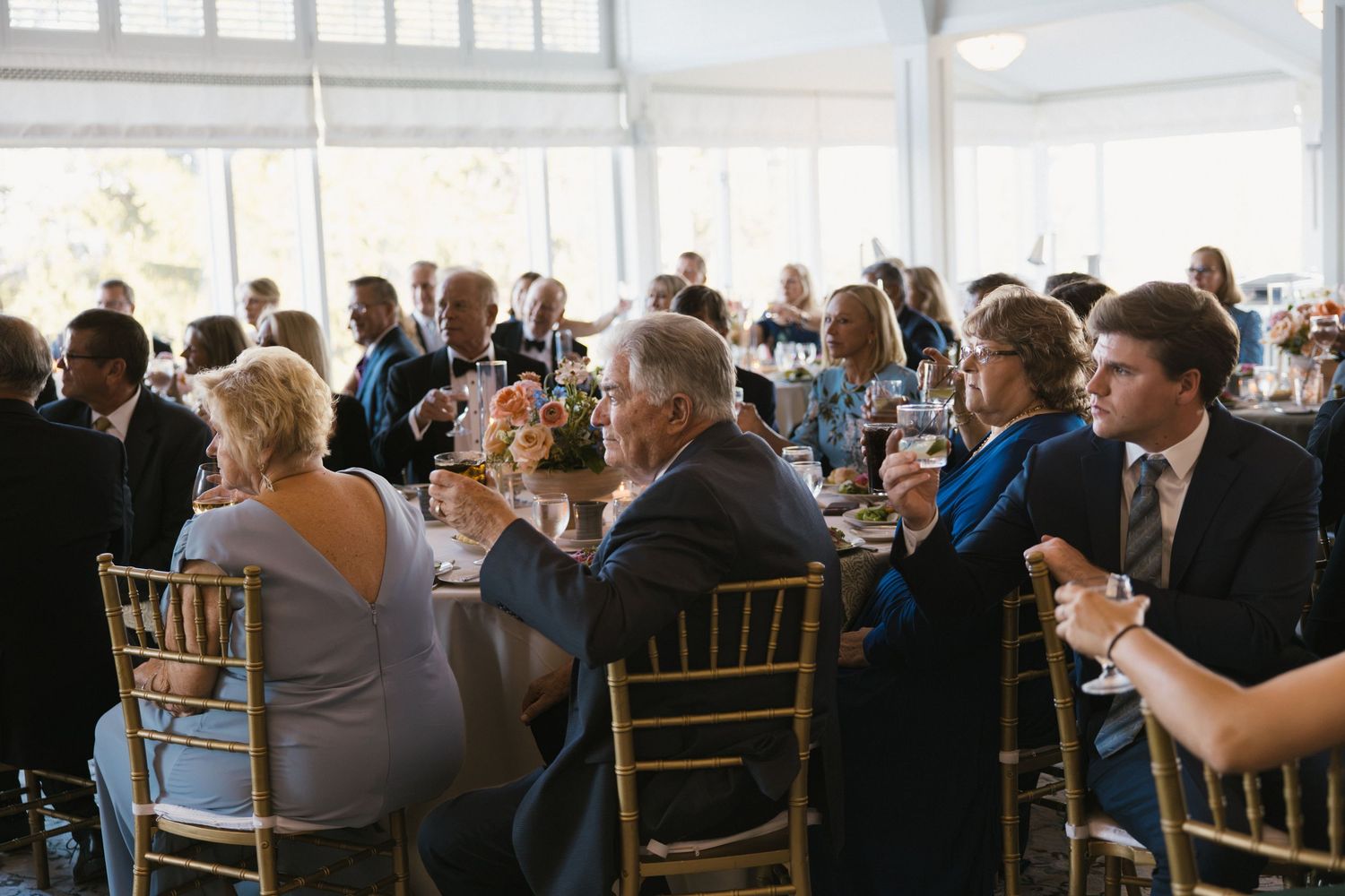 Wedding reception guests seated at round tables with gold chairs raising glasses in a toast.