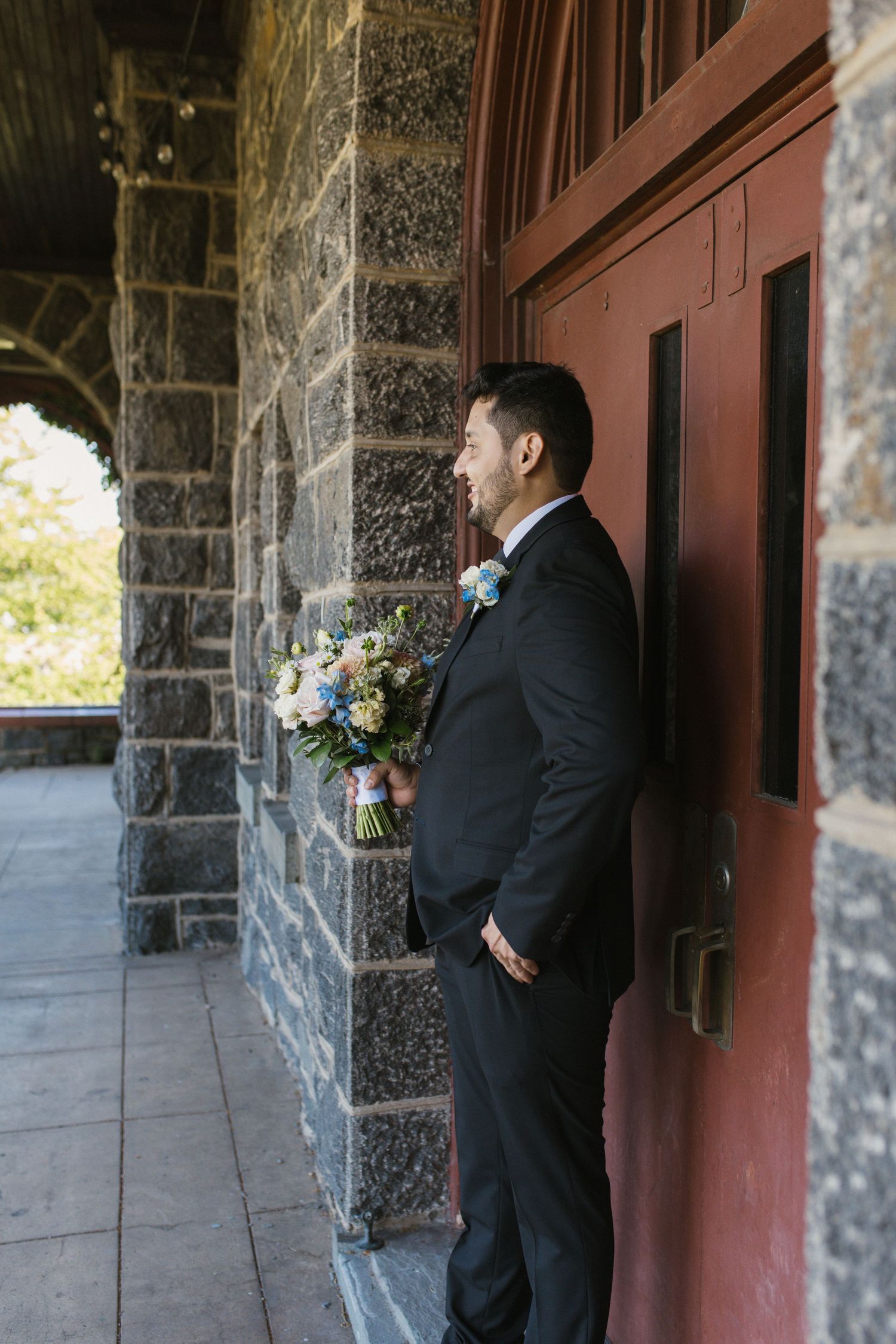 Groom in black tuxedo holding floral bouquet standing by stone church red door.