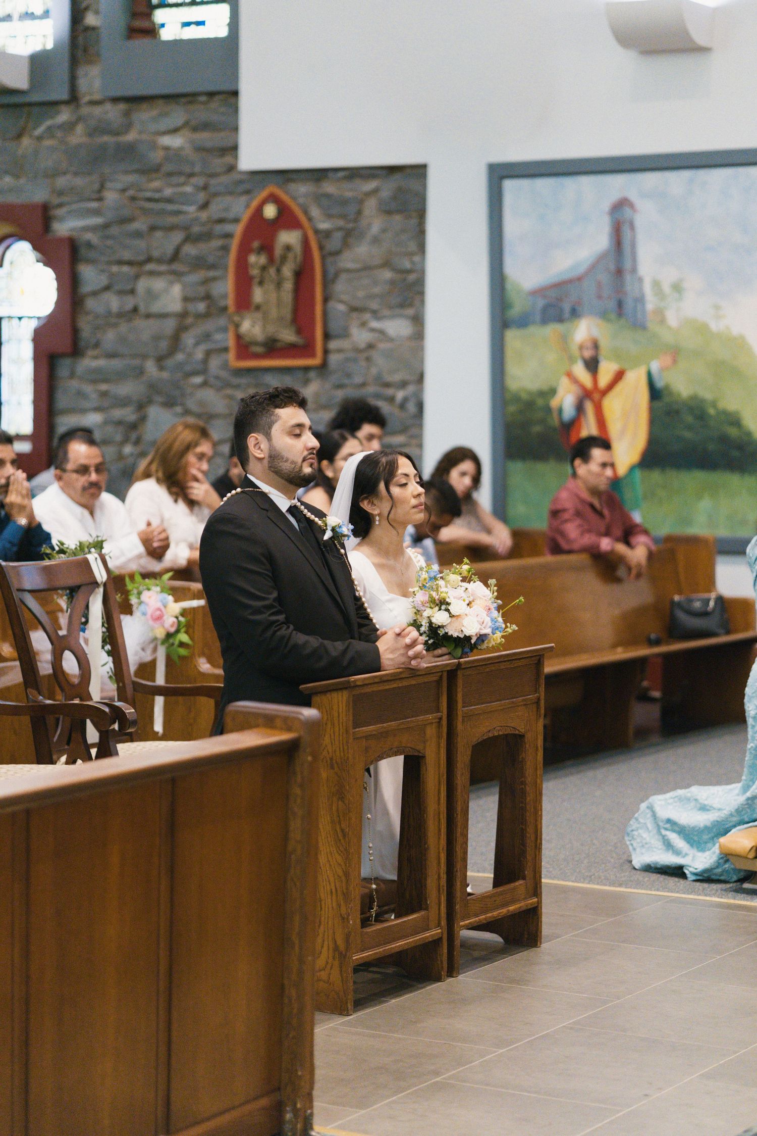 Bride and groom sit attentively in church pew during wedding ceremony with guests in background.
