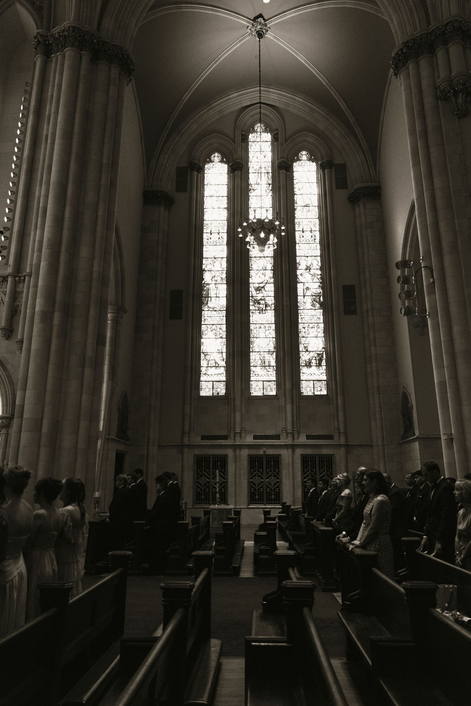 Black and white photo of wedding guests seated in church pews beneath grand stained glass windows.