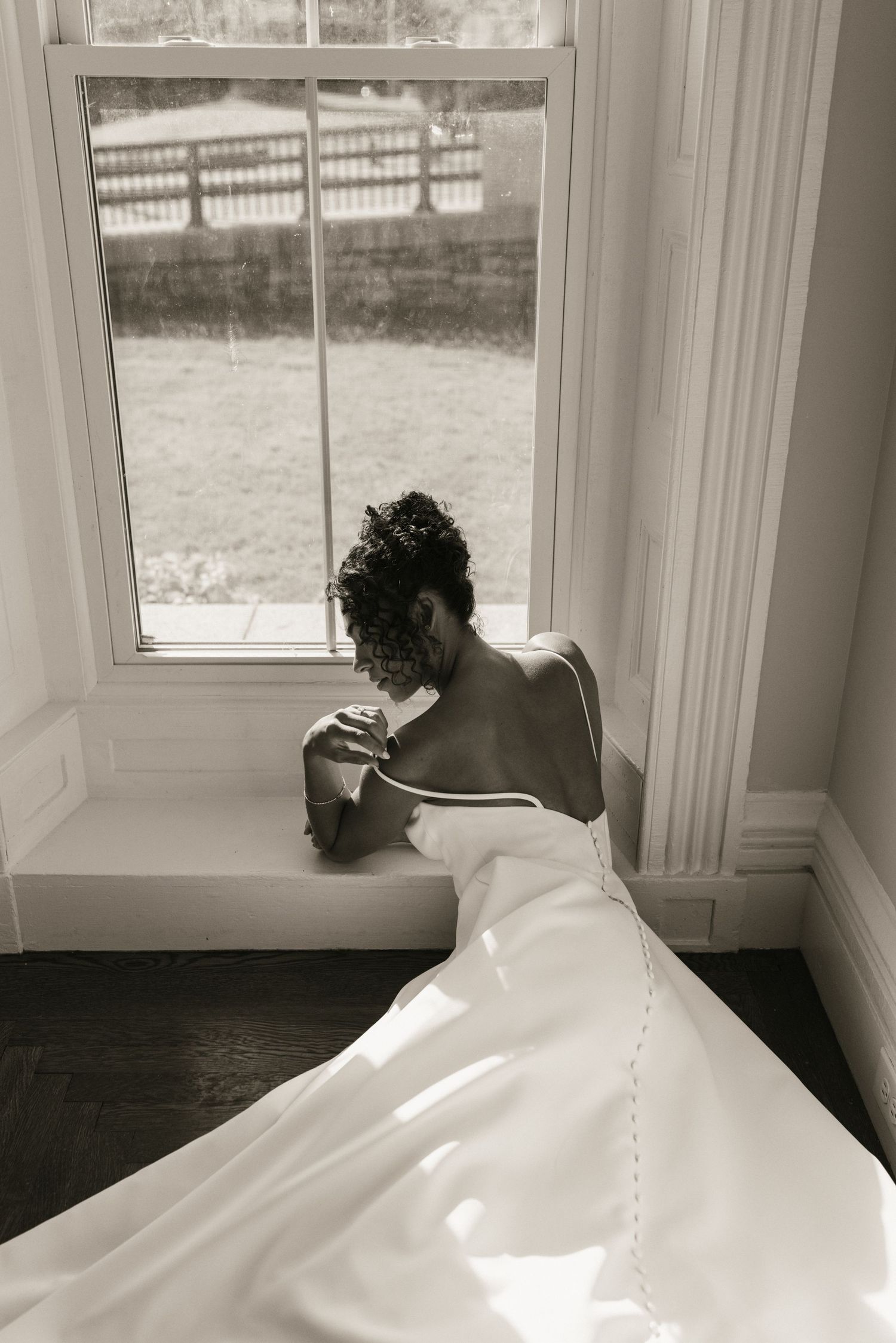 Black and white photo of bride in backless satin gown sitting by window with flowing train.