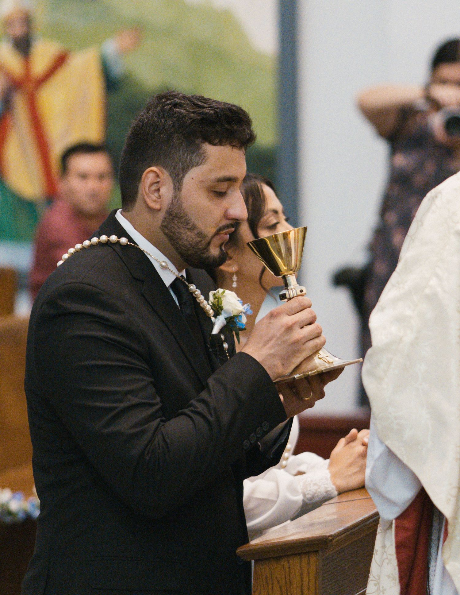 Groom in black suit drinks from golden chalice during Catholic wedding ceremony communion at church altar.