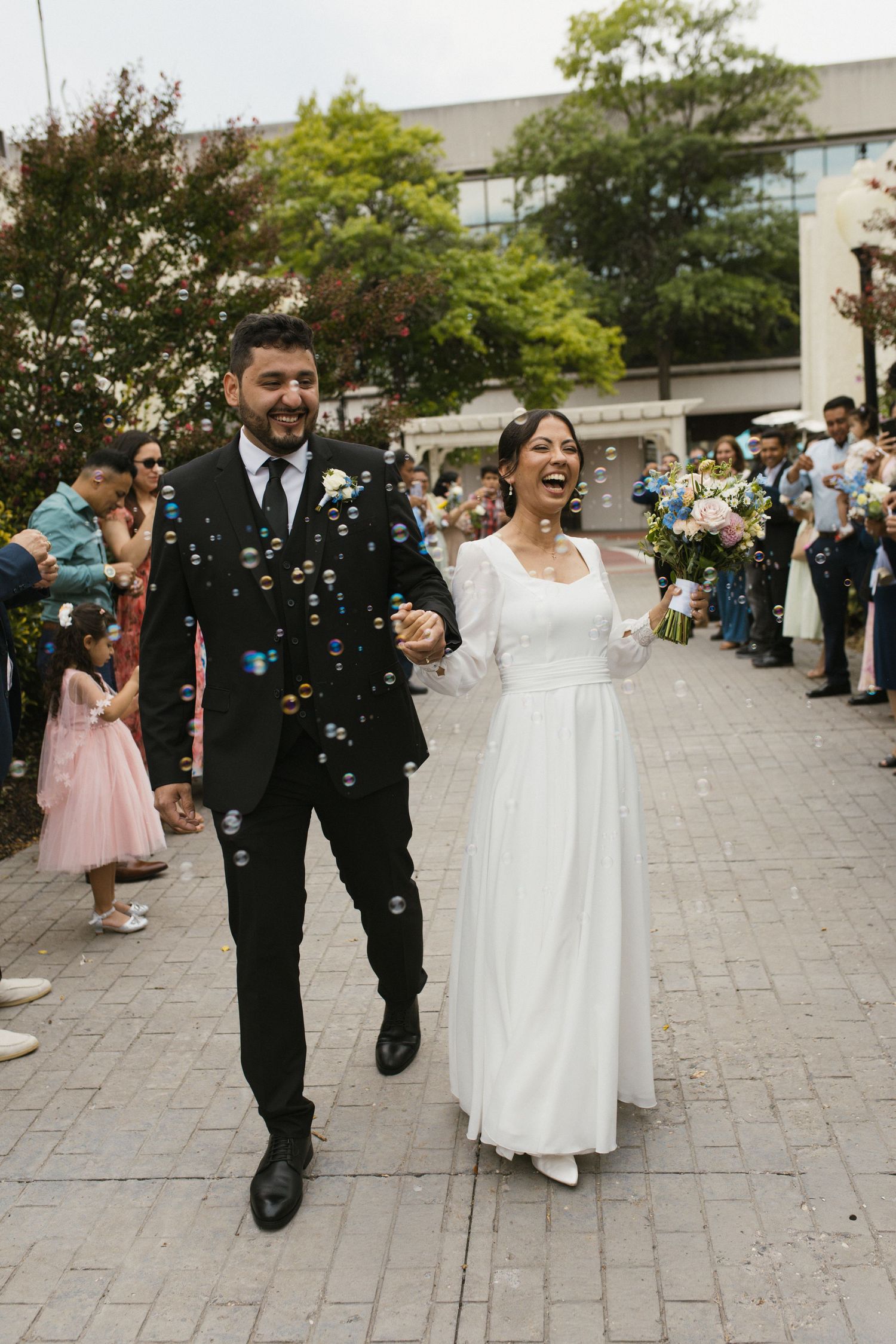 Laughing bride and groom celebrate with bubbles during joyful outdoor wedding reception sendoff.