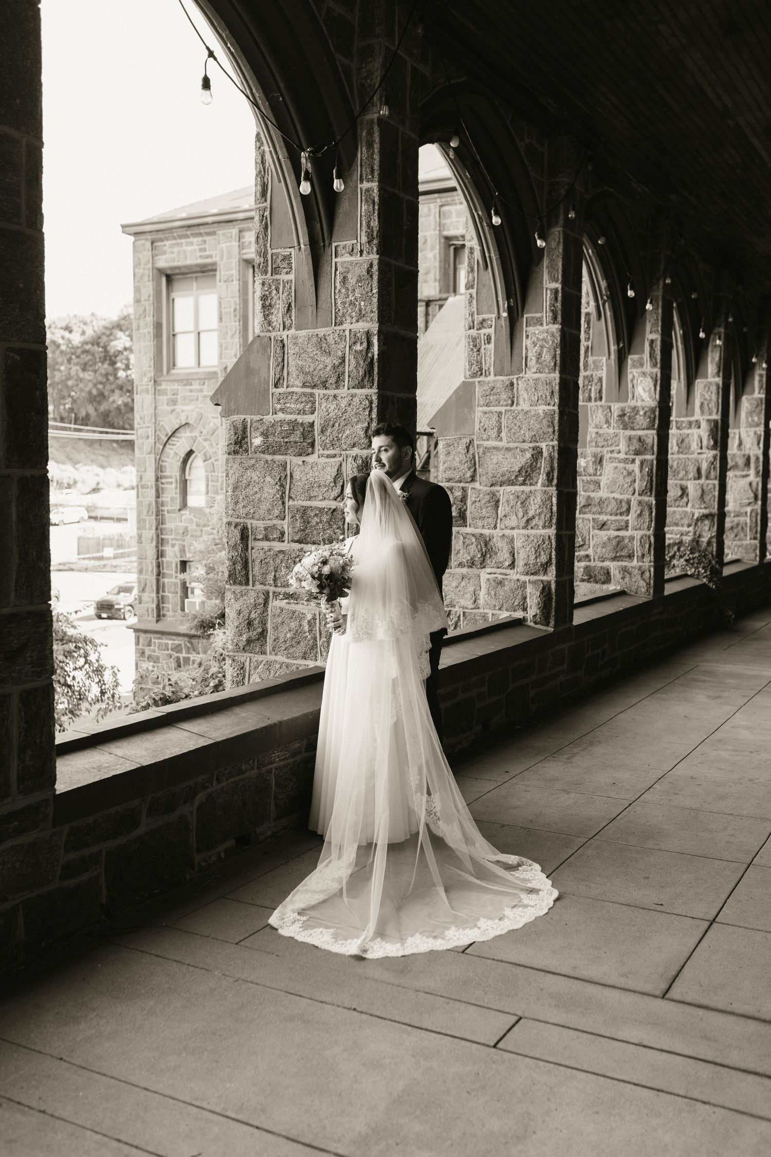 Black and white photo of bride in cathedral veil standing in stone colonnade with groom.