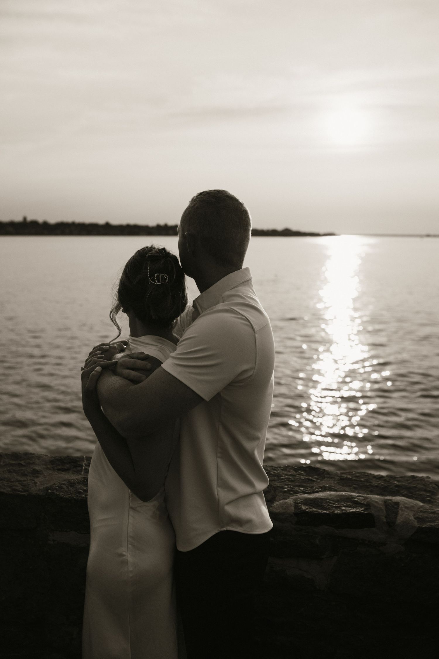 Black and white romantic couple embracing at sunset by the ocean with glowing sun reflection.