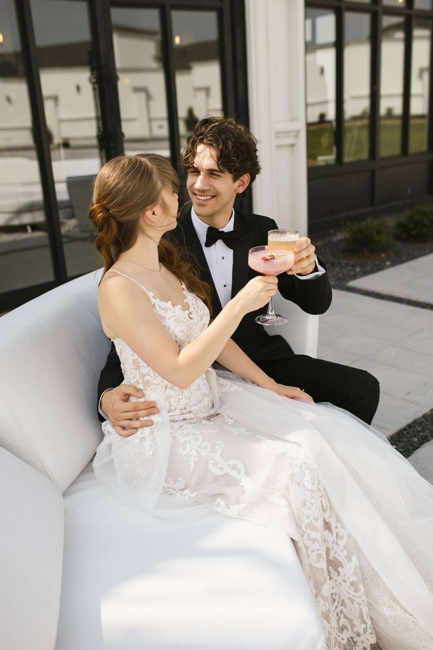 Happy bride and groom in lace wedding attire toasting with cocktails on white outdoor lounge furniture.