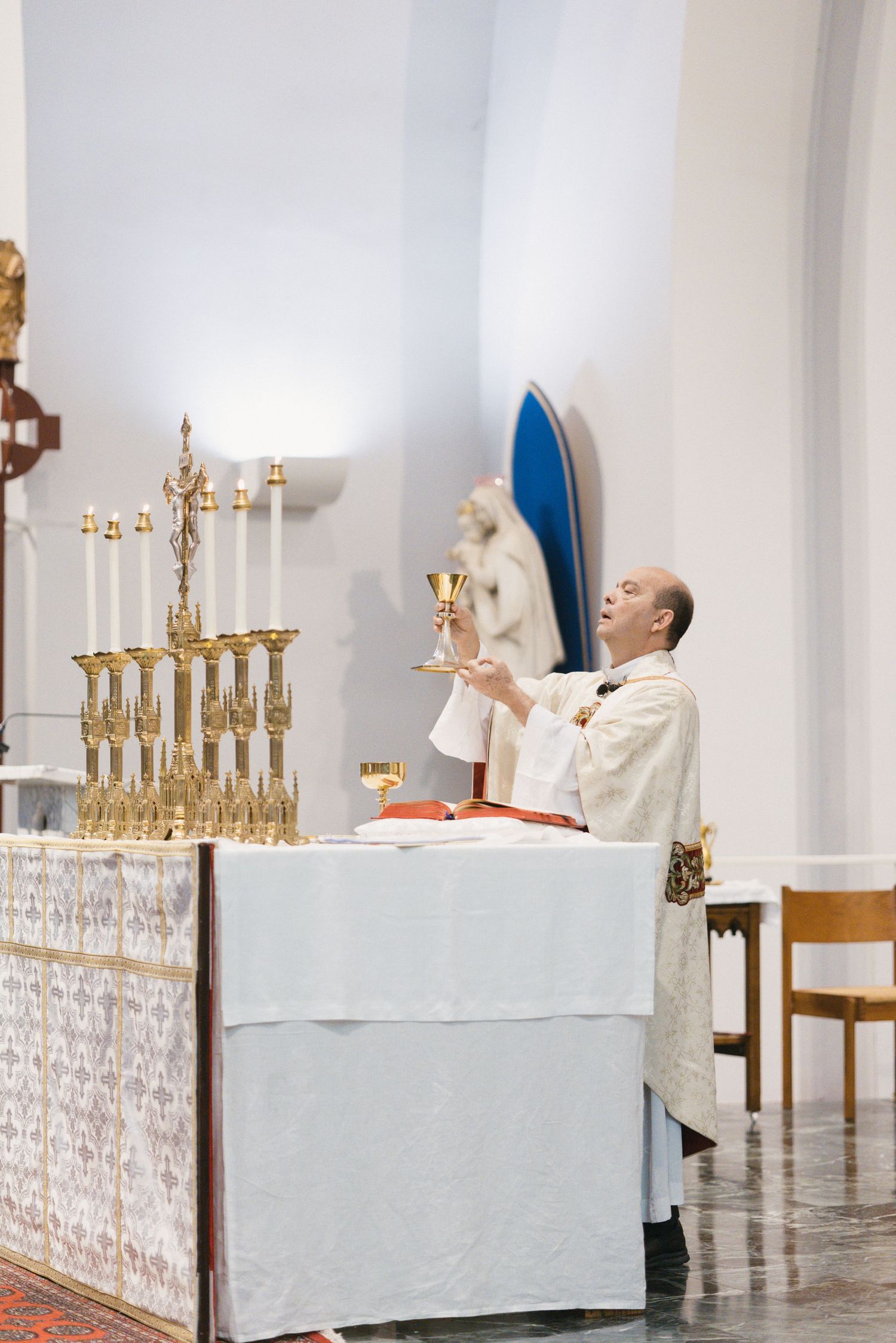 Catholic priest elevates golden chalice during wedding Mass at ornate altar adorned with candelabras and white linens.