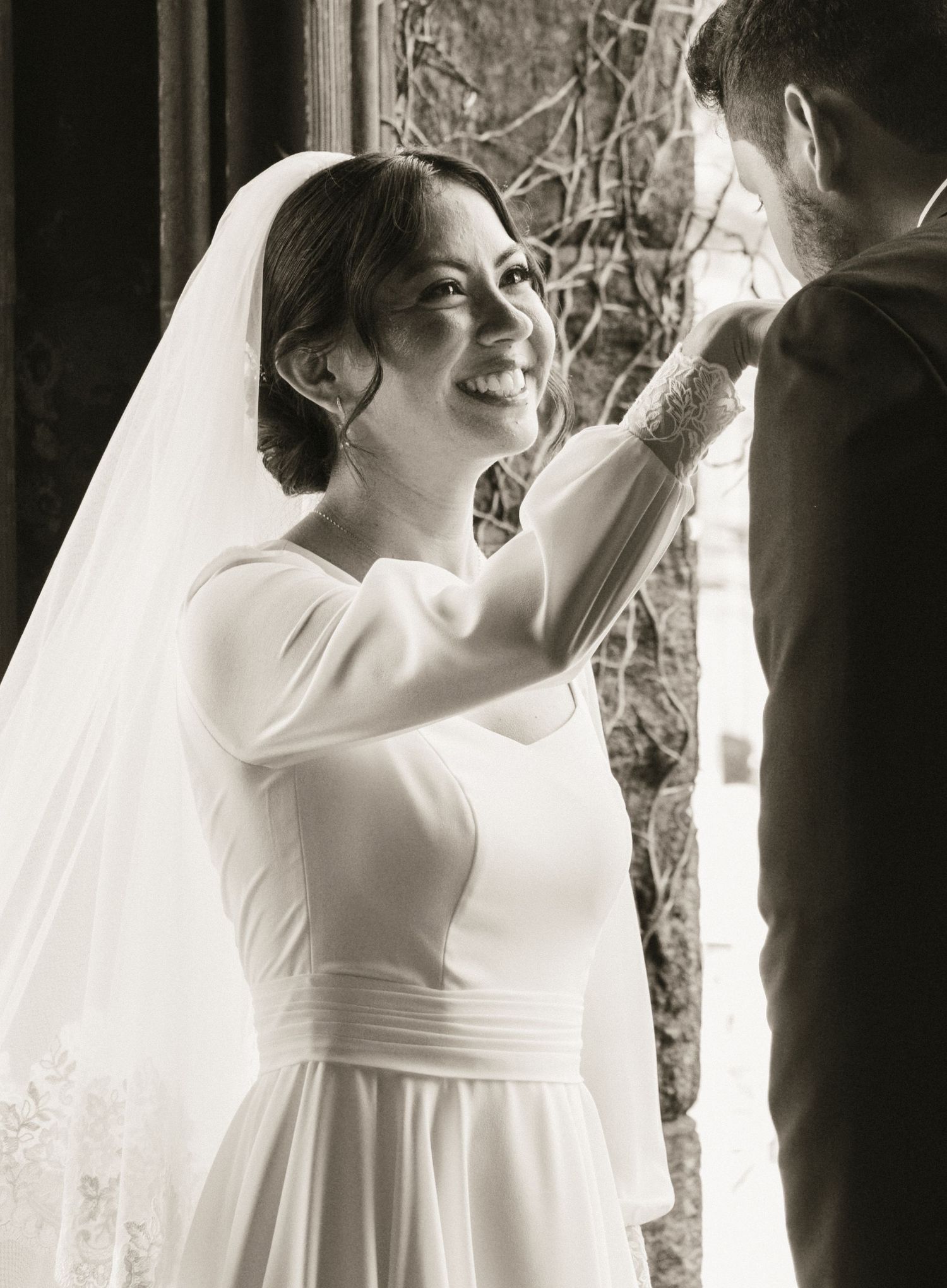 Black and white photo of a smiling bride in a white gown and veil adjusting the groom's suit lapel.