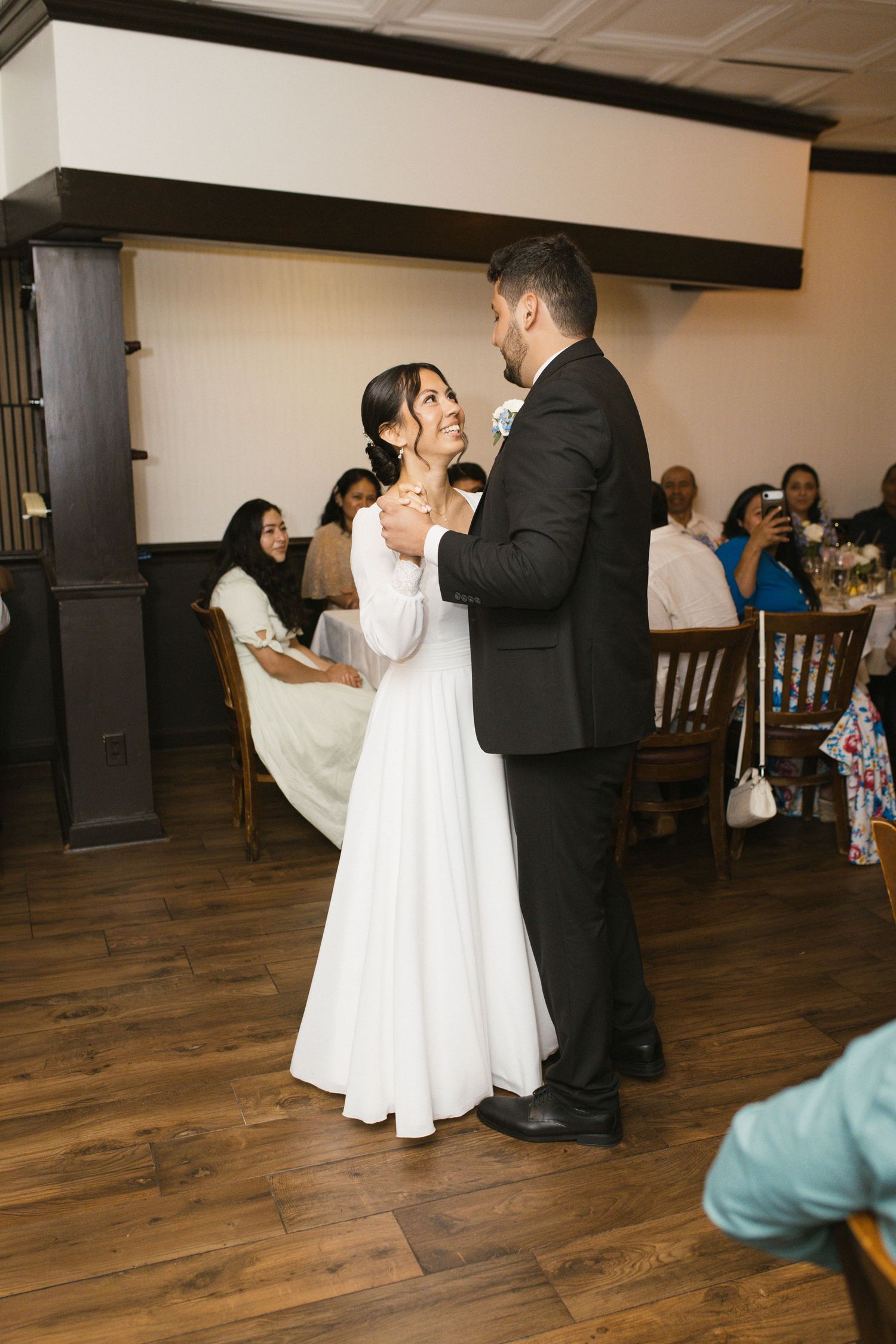 Bride in elegant white gown smiling at groom during first dance at wedding reception.