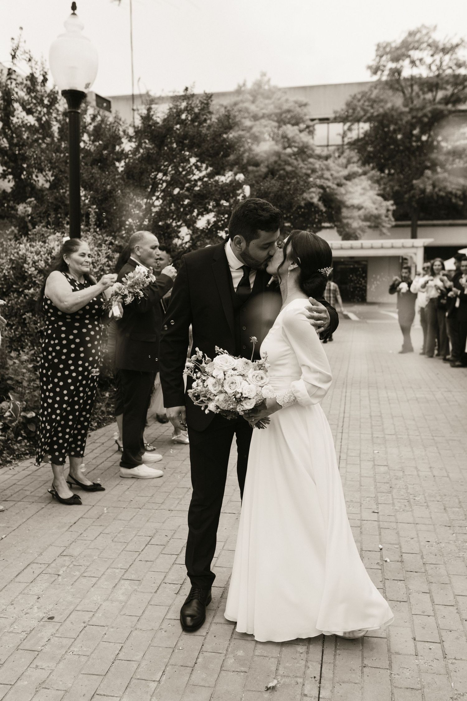 Black and white photo of a bride and groom sharing a kiss outdoors while guests celebrate around them.