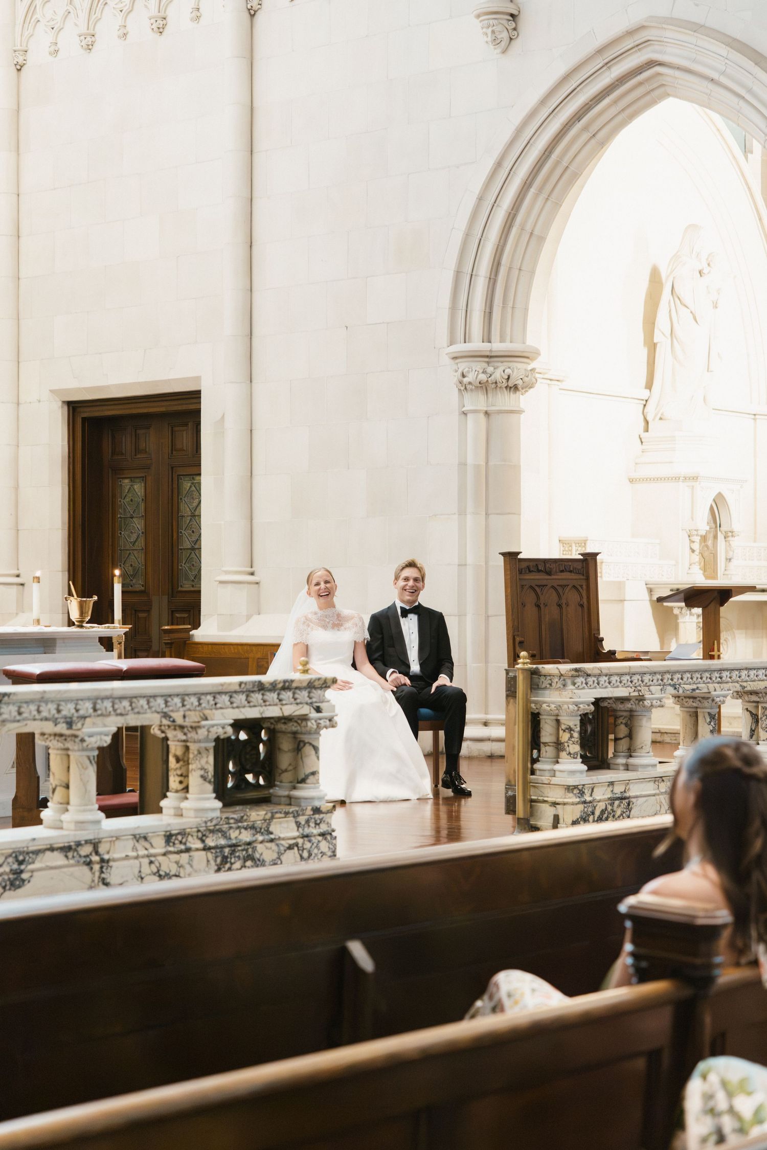 Bride and groom seated at church altar during wedding ceremony, surrounded by elegant marble architecture.