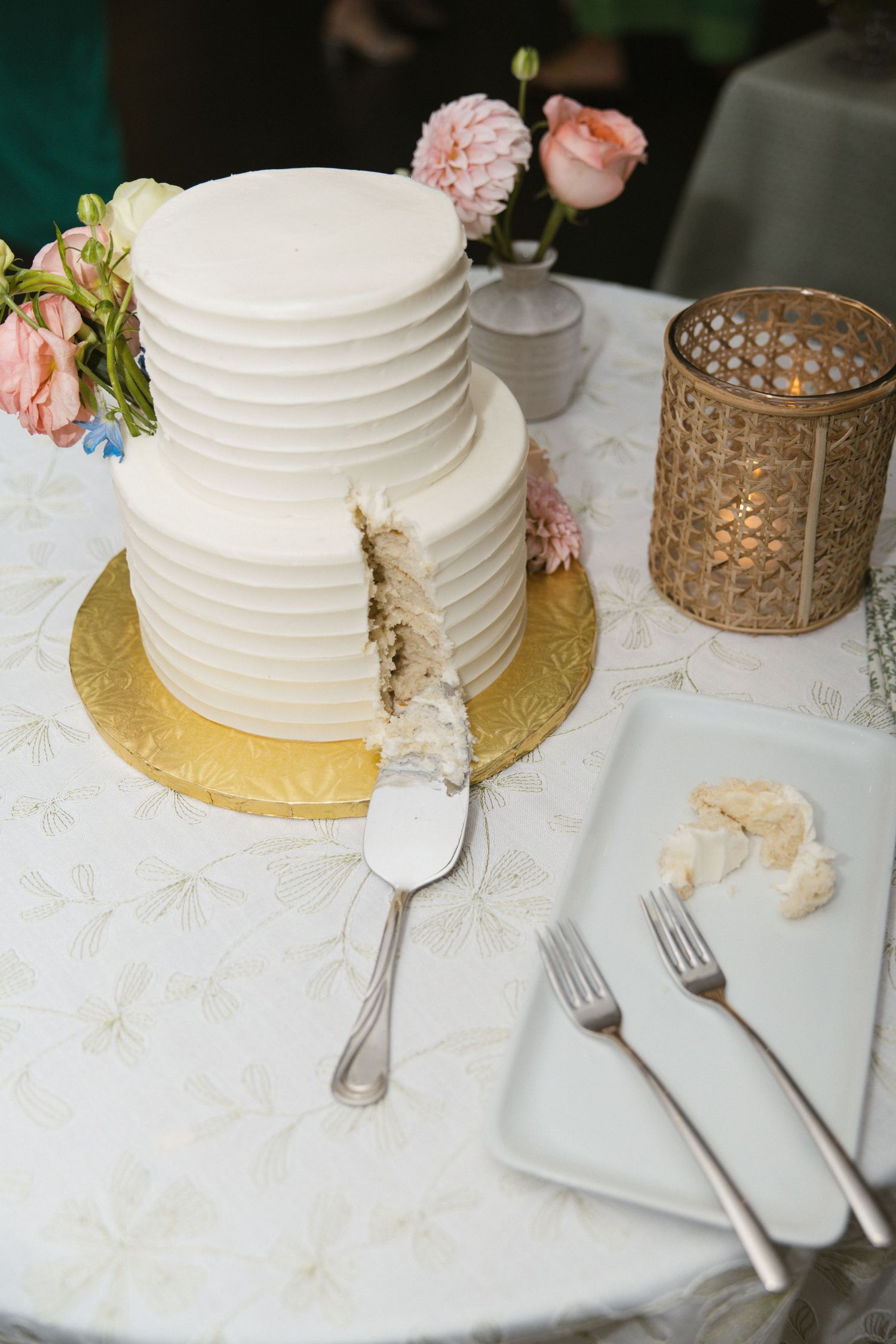 Two-tier white ribbed wedding cake on gold stand with pink flowers, partially sliced, beside forks and candle.