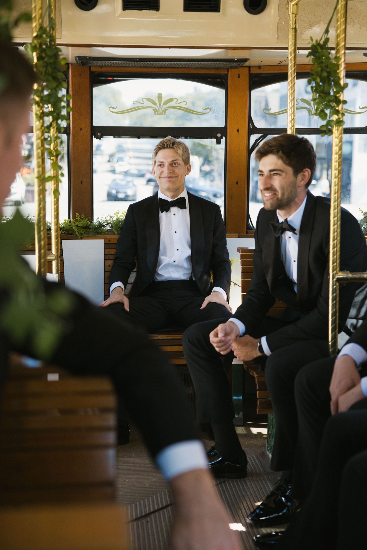 Two grooms in black tuxedos smile while riding a decorated trolley car on their wedding day.