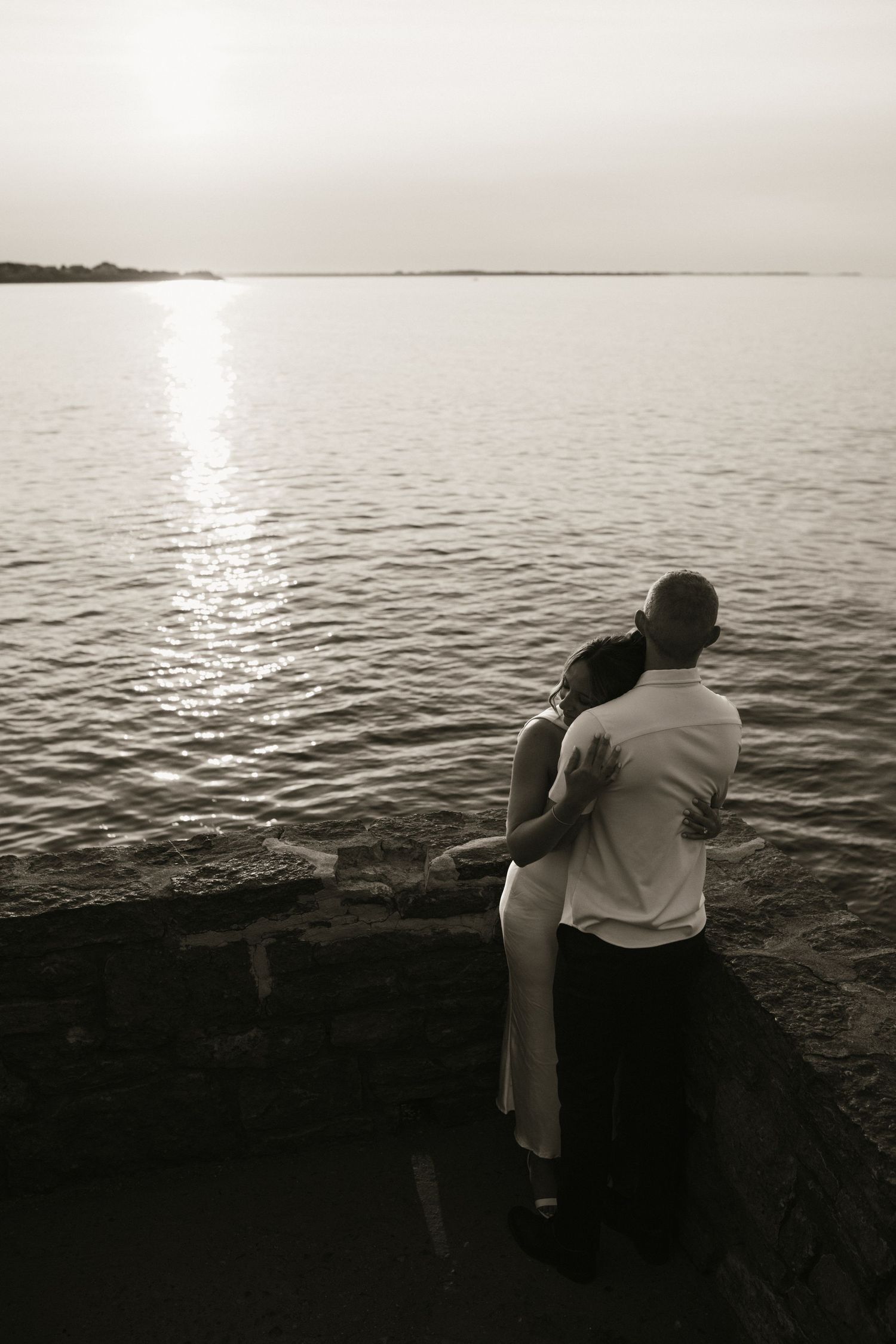 Black and white photo of couple embracing while gazing at a serene waterfront sunset.