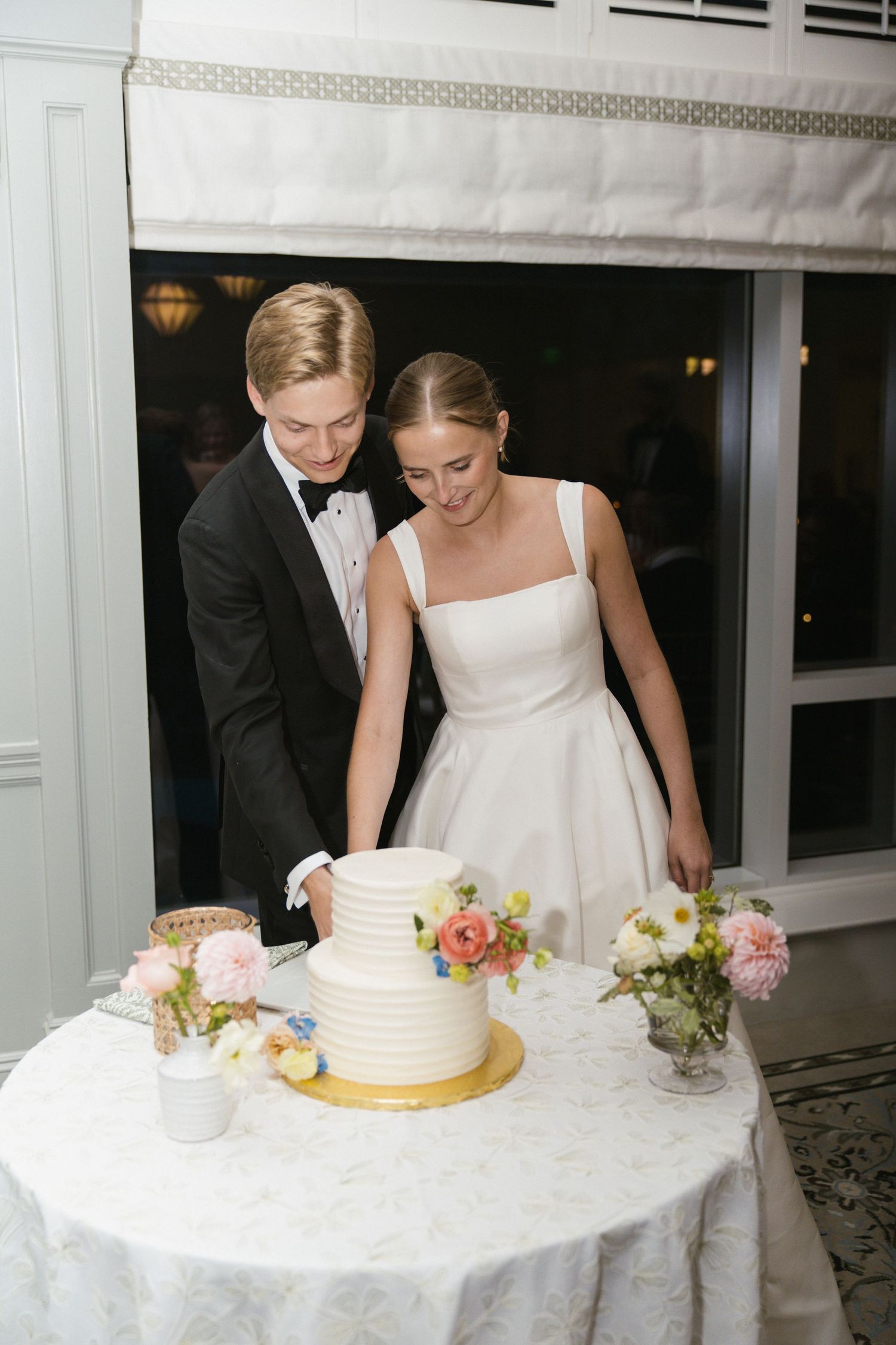 Smiling bride and groom cut their elegant three-tier white wedding cake adorned with colorful flowers.