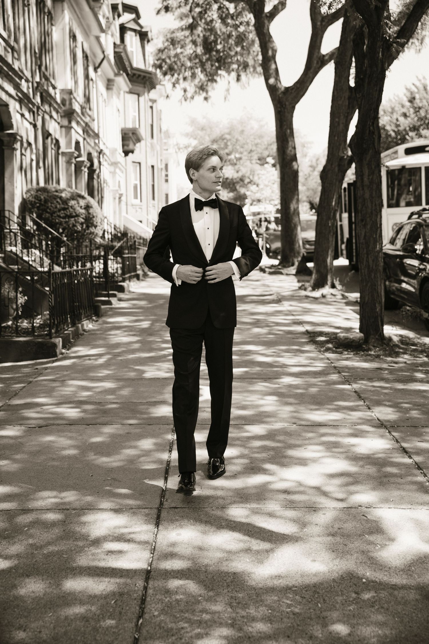 A young man in a black tuxedo and bow tie walks confidently down a tree-lined urban sidewalk in a black and white photo.