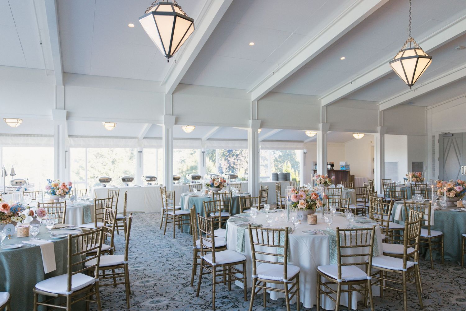 Elegant wedding reception hall with gold chiavari chairs, blue linens, floral centerpieces, and vaulted white ceilings.