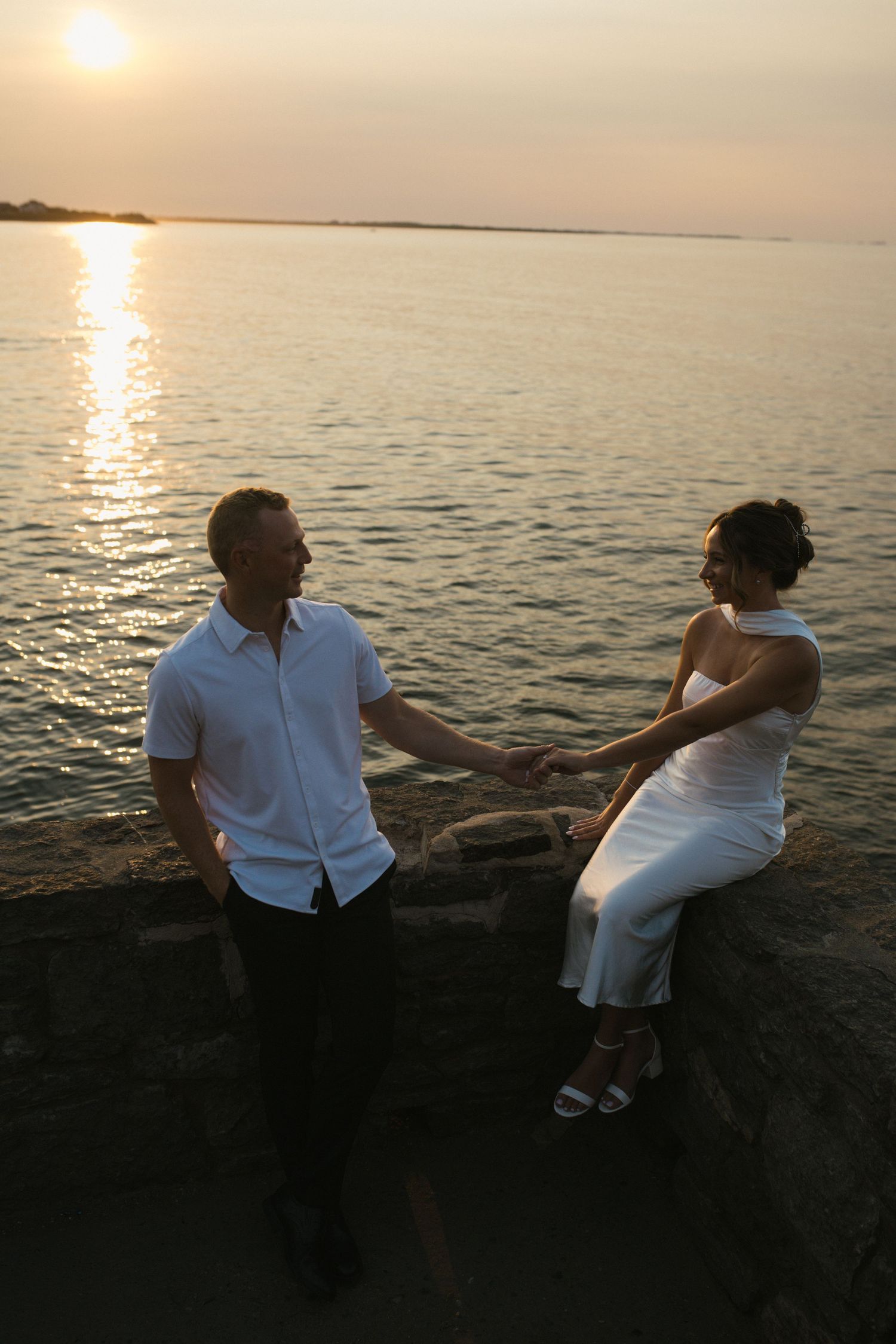 Couple holding hands and smiling at each other by the ocean during a golden sunset on stone wall.
