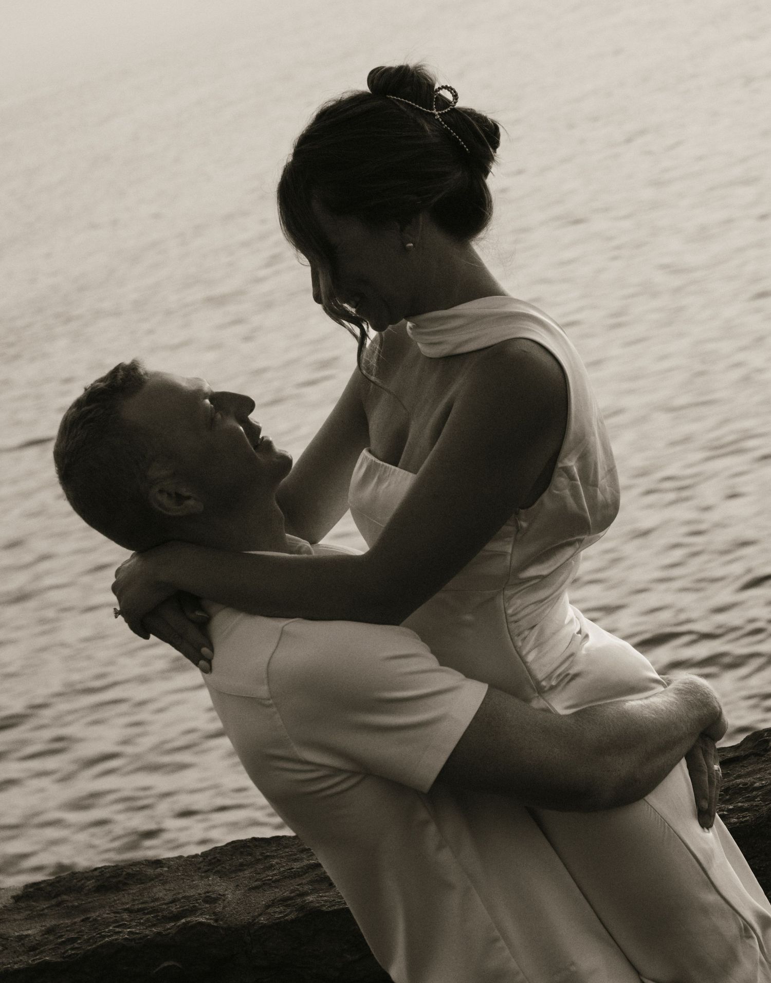 A couple in white wedding attire share a romantic embrace by the water's edge in a black and white photo.