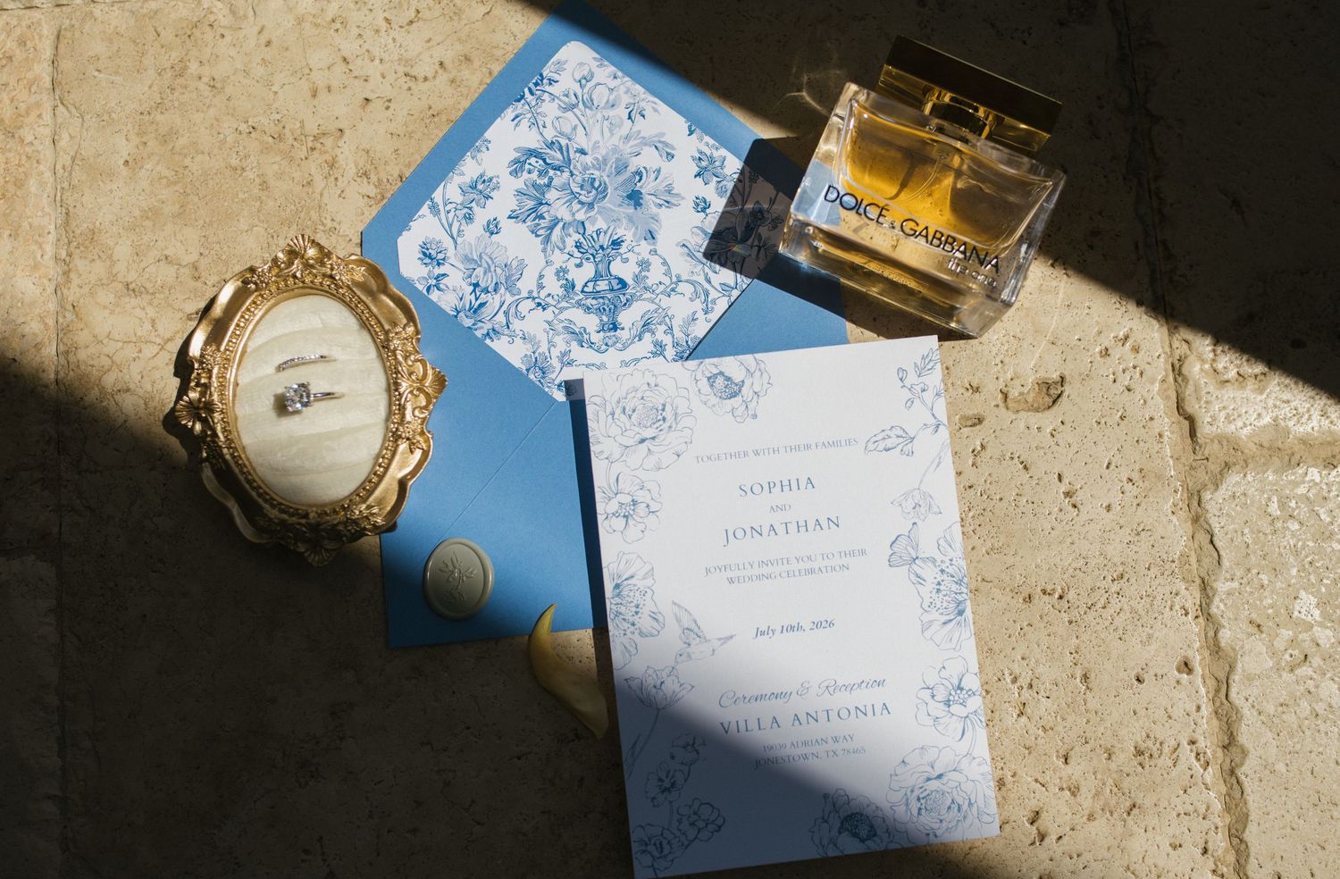 Blue wedding stationery with floral envelope liner, gold frame ring holder, and Dolce & Gabbana perfume on stone surface.