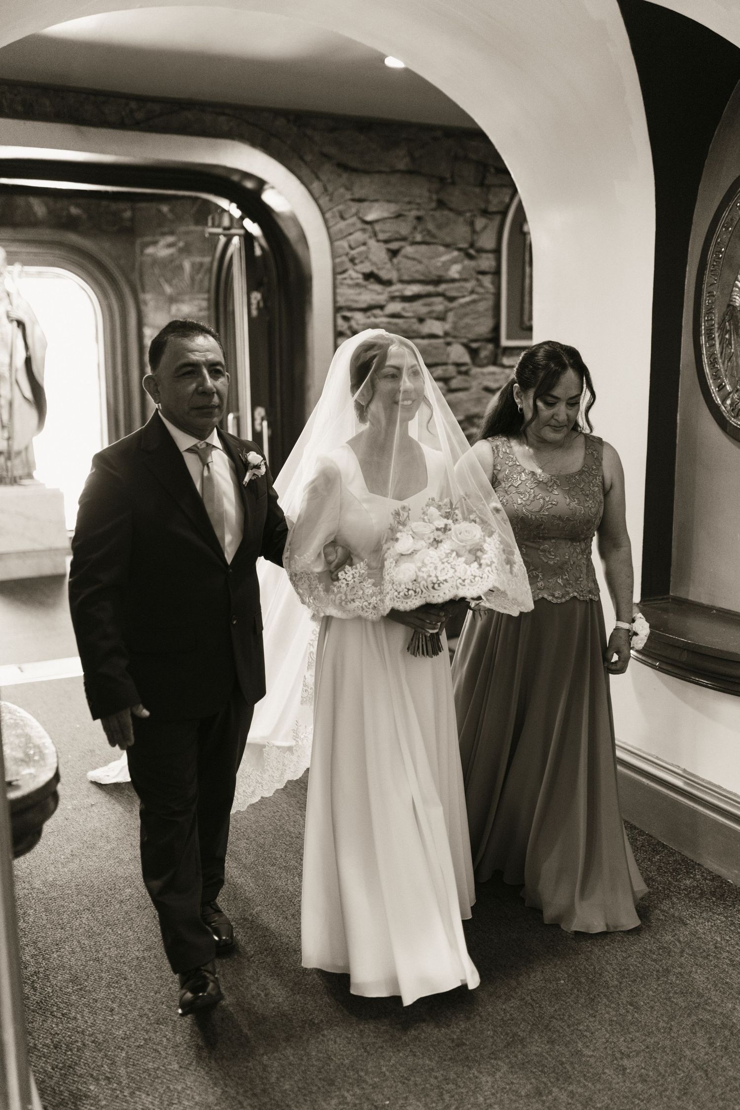 Black and white photo of bride walking down church aisle with parents holding bouquet.