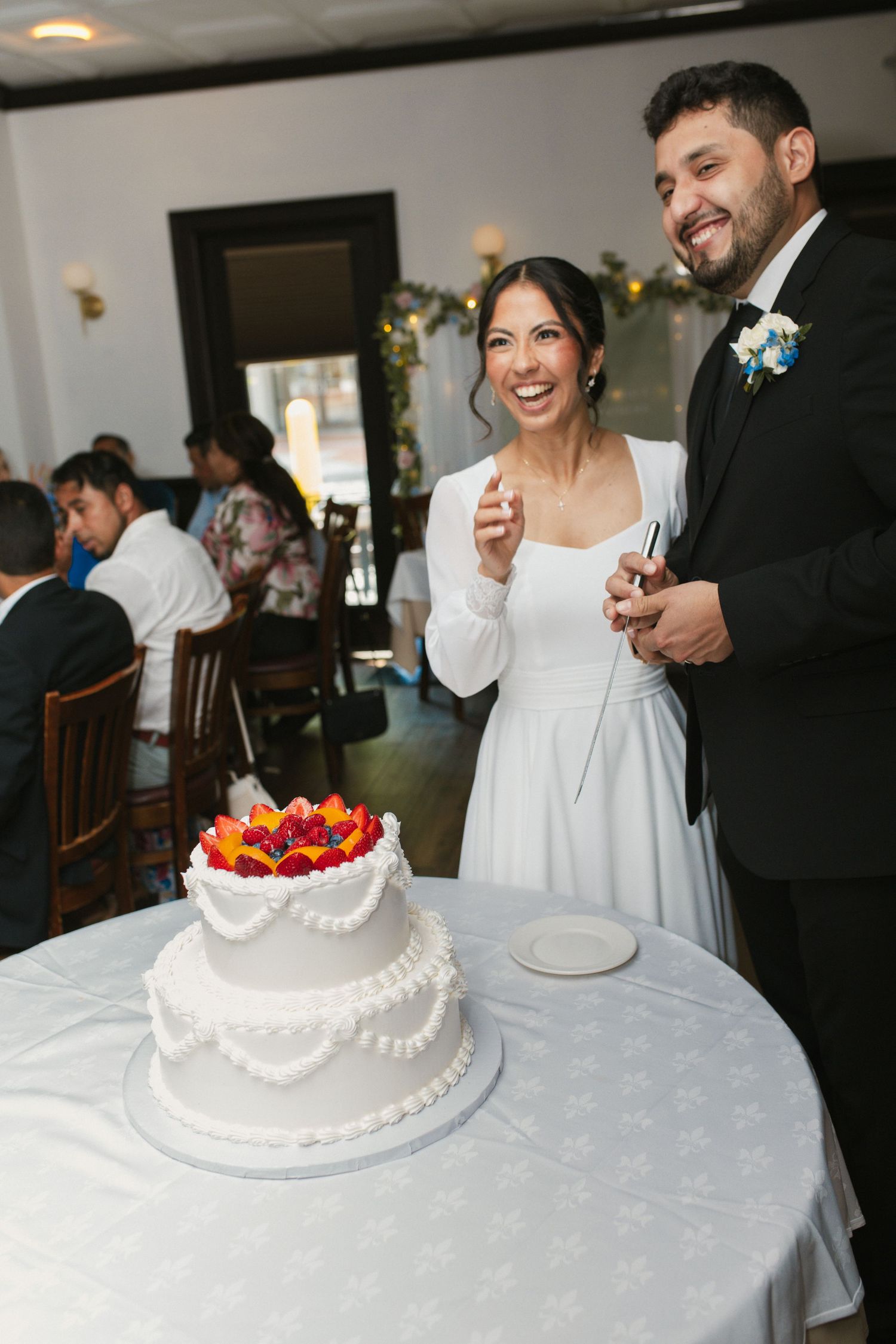 Smiling bride and groom cutting their two-tier white wedding cake with colorful floral topper.