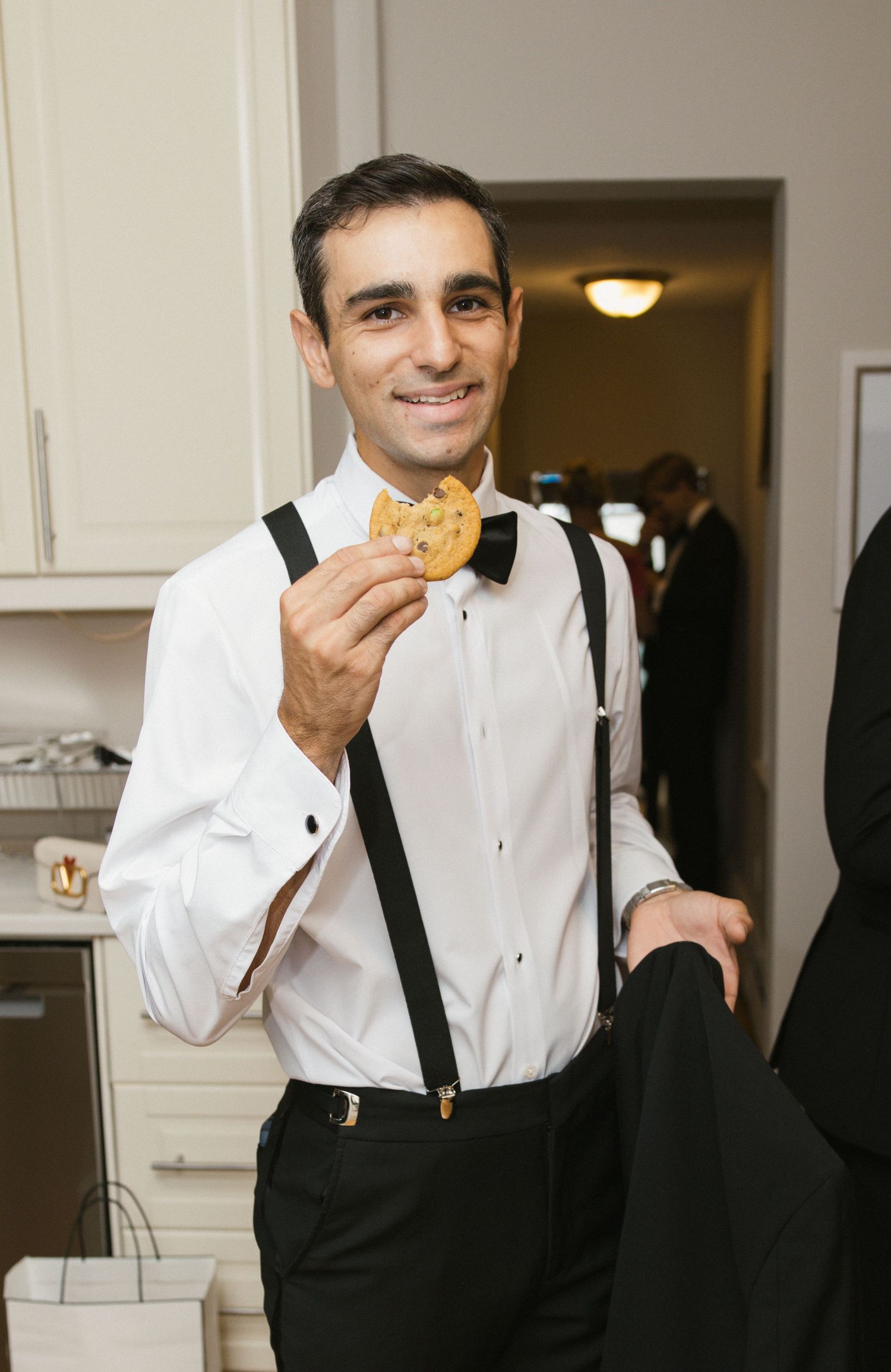 Smiling man in white dress shirt with suspenders and bow tie holding a chocolate chip cookie at a formal event.