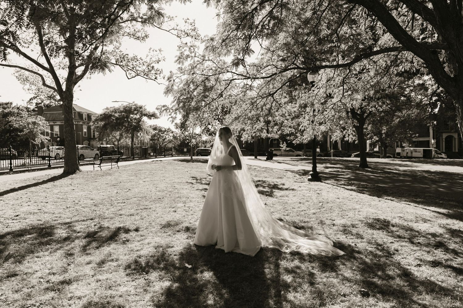 Black and white photo of a bride in a flowing wedding gown standing in a sunlit park surrounded by trees.